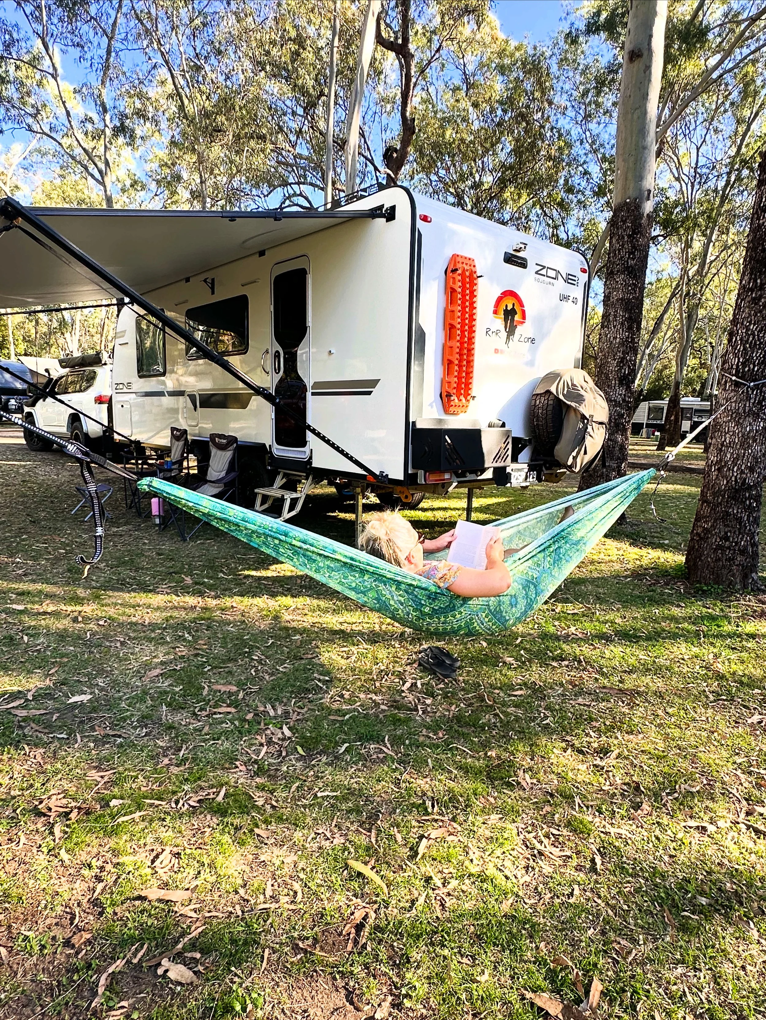 Child lying in a green hammock reading a book next to a white travel trailer parked in a wooded area.