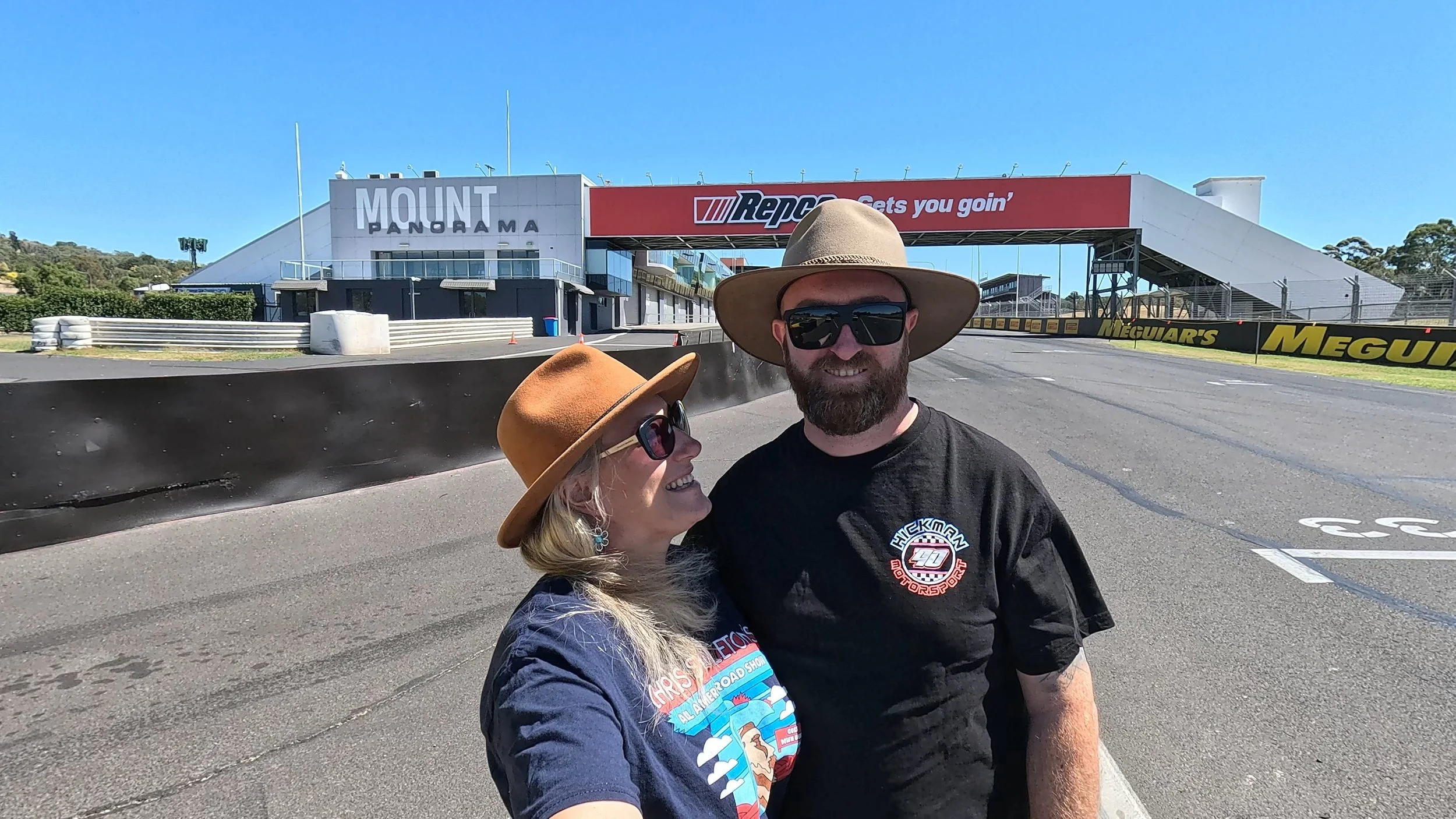 A smiling woman and man standing on a race track at Mount Panorama, with a large building and banners in the background, both wearing wide-brimmed hats and sunglasses.