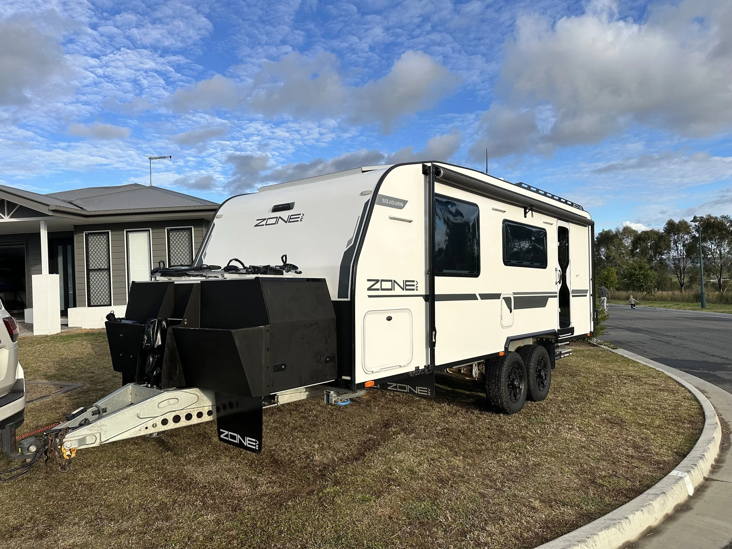 A white caravan with black accents labeled 'ZONE' parked on a residential street on a sunny day with a partly cloudy sky. The trailer is attached to a vehicle and has a black utility box at the front.