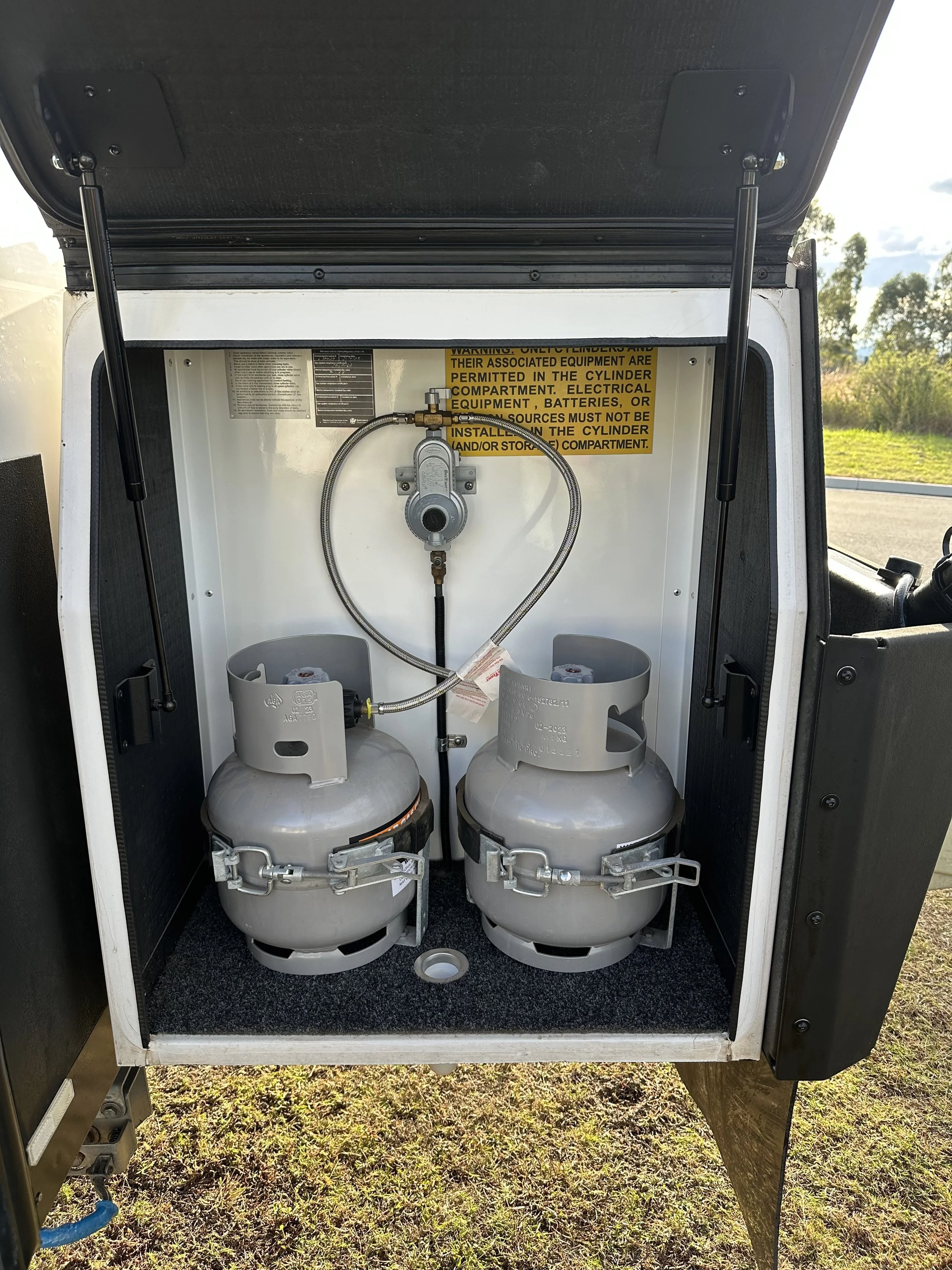 An outdoor gas compressor with two large cylinders, a pressure gauge, and a warning label, housed in a white metal enclosure with an open door, on grass with trees and a cloudy sky in the background.