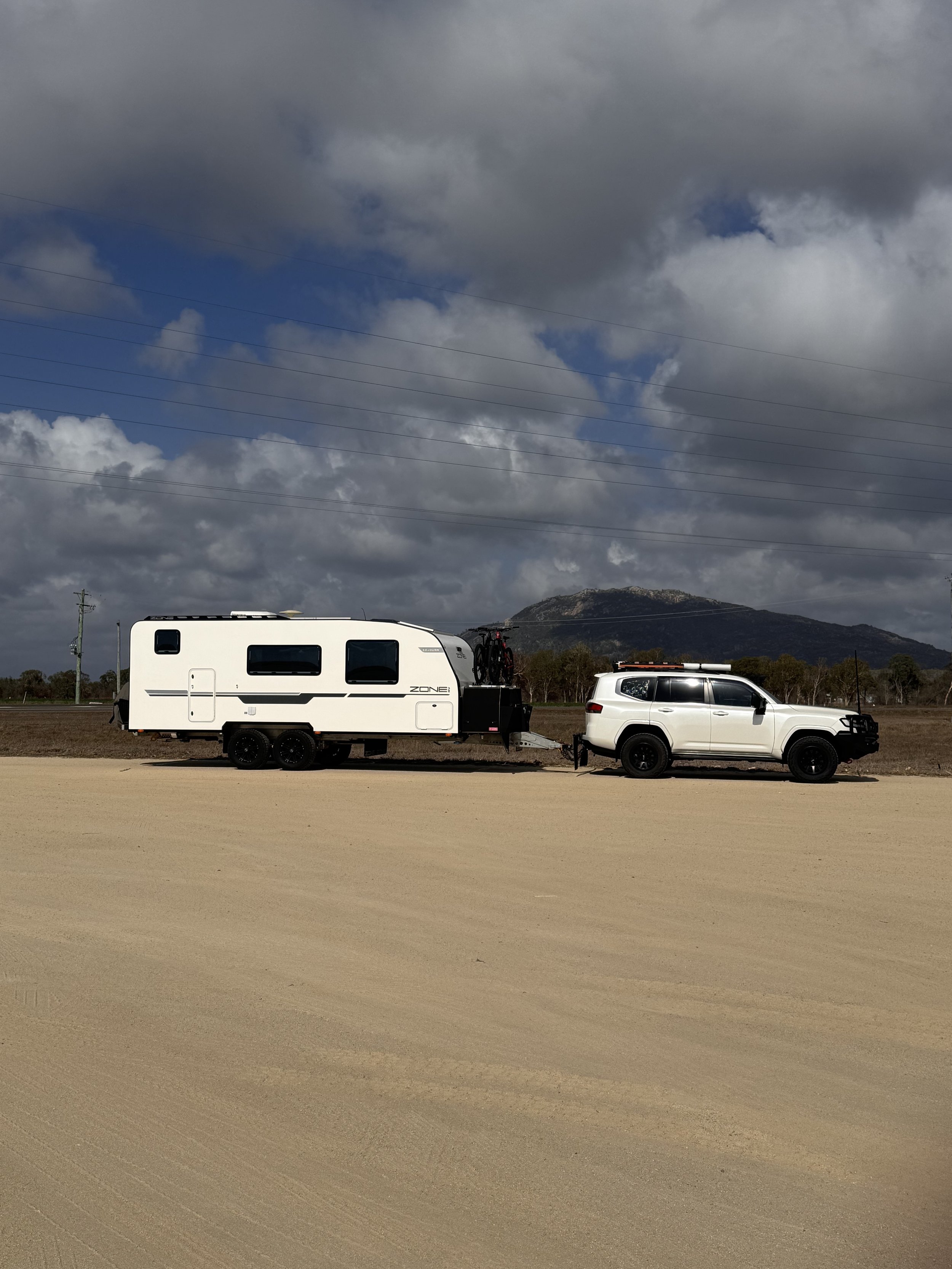 A Zone RV Caravan attached to a white SUV parked on a gravel ground with a mountain and cloudy sky in the background.