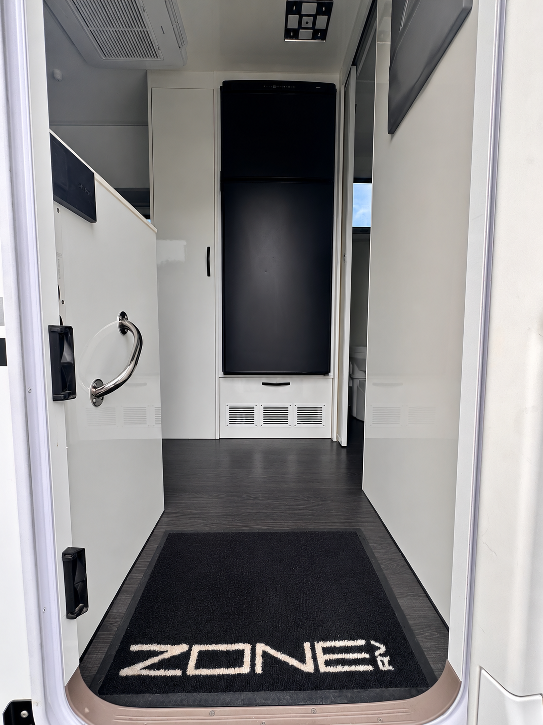 View inside a caravan with a black refrigerator, white cabinets, a small bathroom door, and a black mat with 'ZONE RV' written on it.