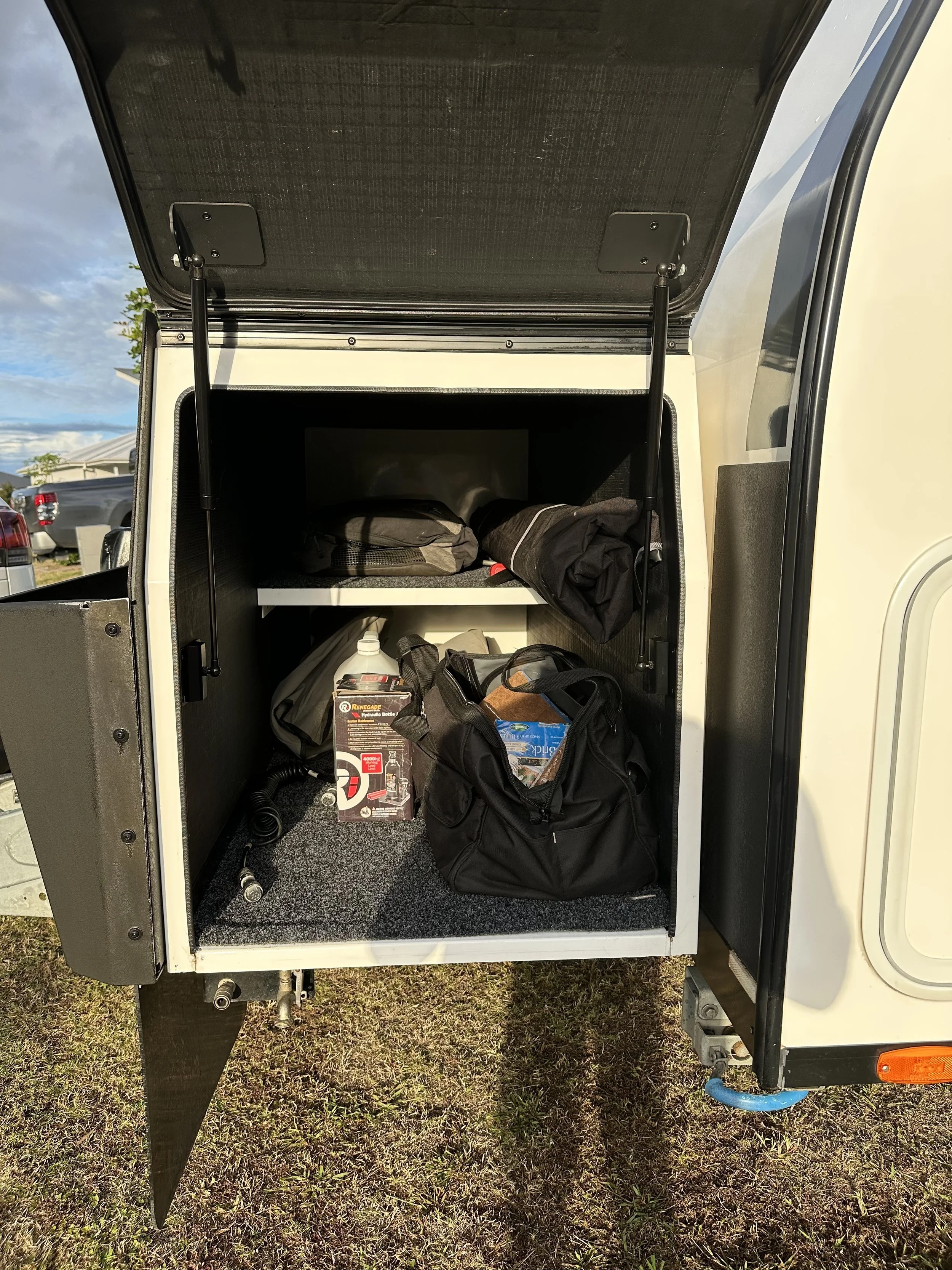 Open caravan storage area with bags, supplies, and a box with a hose. The ground is grassy and the sky is partly cloudy.