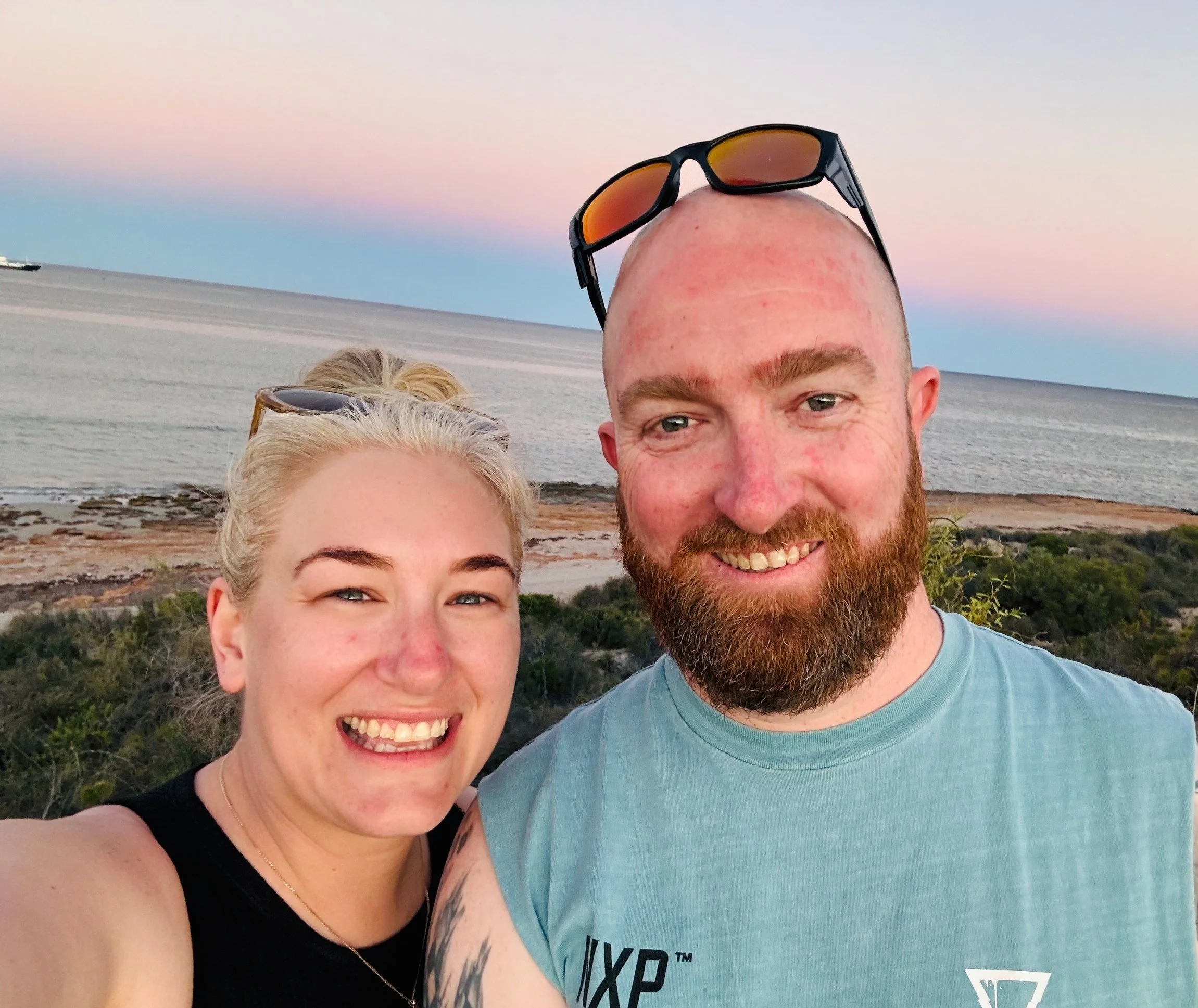 A smiling man and woman taking a selfie outdoors near the beach during sunset. The man has a beard and sunglasses on his bald head, and the woman has blonde hair, sunglasses on her head, and is wearing a black top. They are standing on a grassy area with the ocean, shoreline, and colorful sky with clouds in the background.