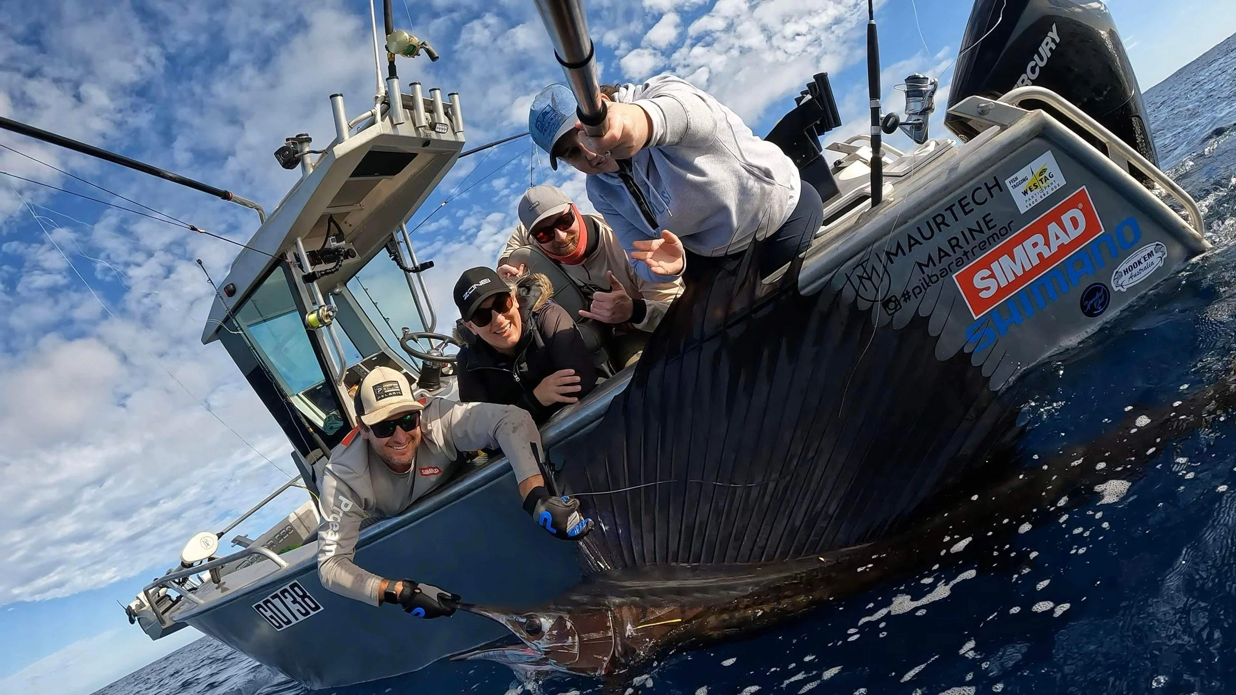 Four people on a boat with fishing gear and a recently caught Sailfish, smiling and taking a selfie over the ocean on a partly cloudy day.