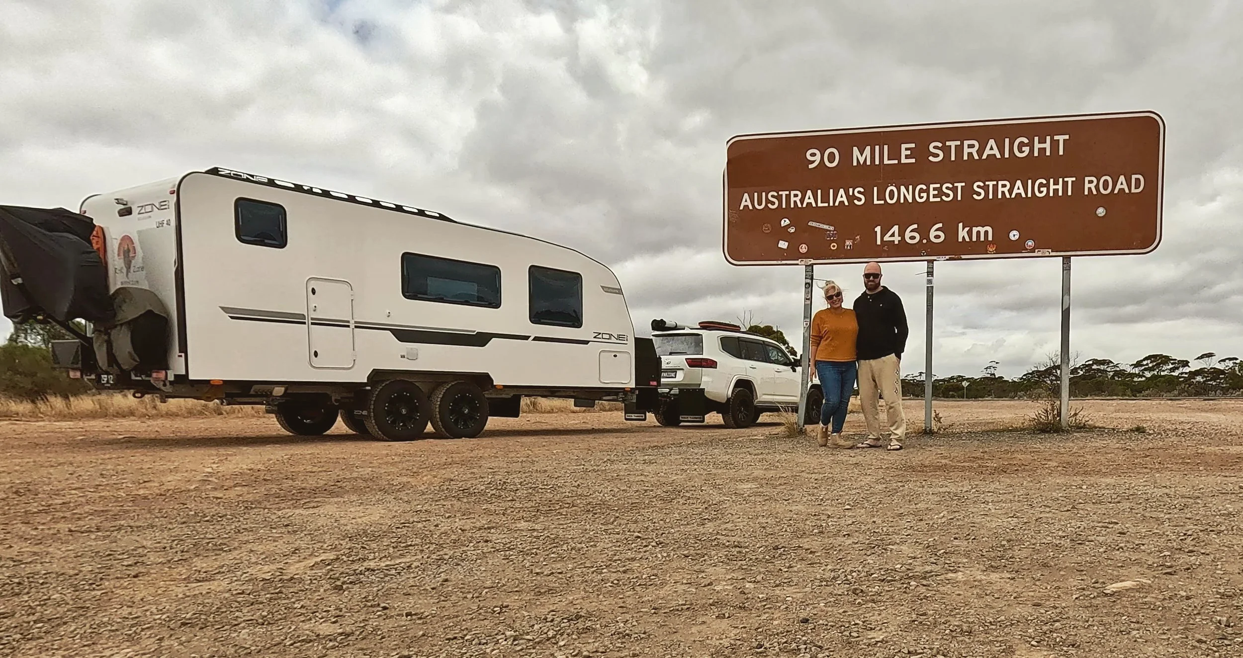 Two people standing next to a large brown sign that reads '90 Mile Straight, Australia's Longest Straight Road, 146.6 km,' with a white vehicle, a caravan, and a white car parked on a dirt road under a cloudy sky.