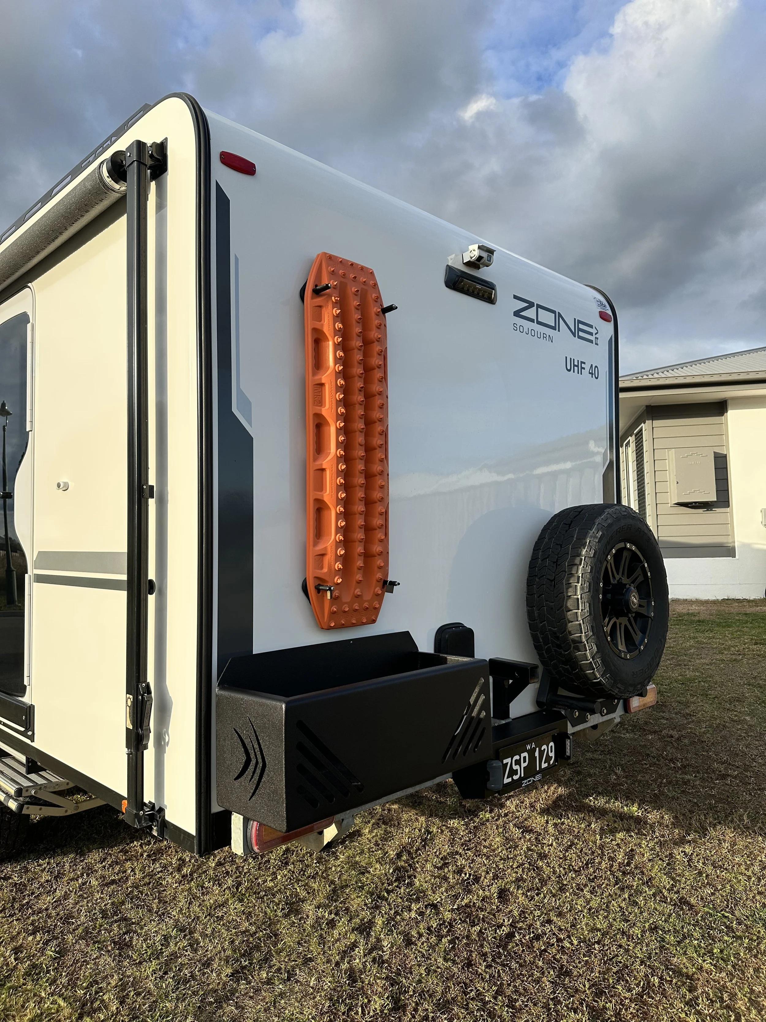 The rear side of a white off-road caravan with a spare tire, orange maxx traxx, and various accessories, parked on a grassy field under cloudy sky.