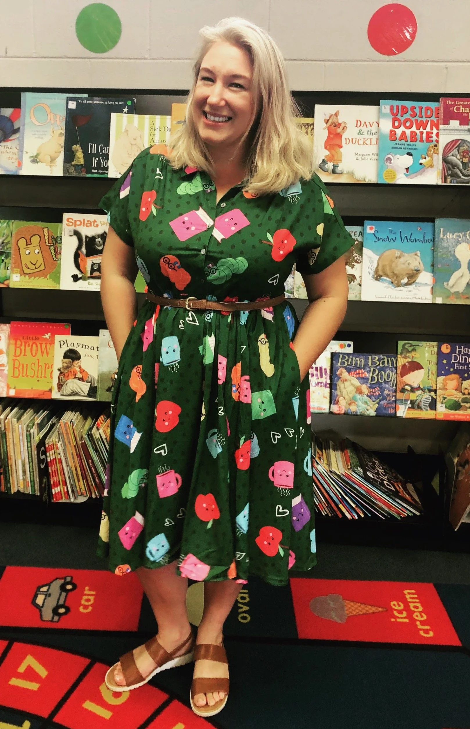 A woman standing in front of a bookshelf filled with children's books, wearing a green dress with a colorful cartoon book pattern, and tan sandals.