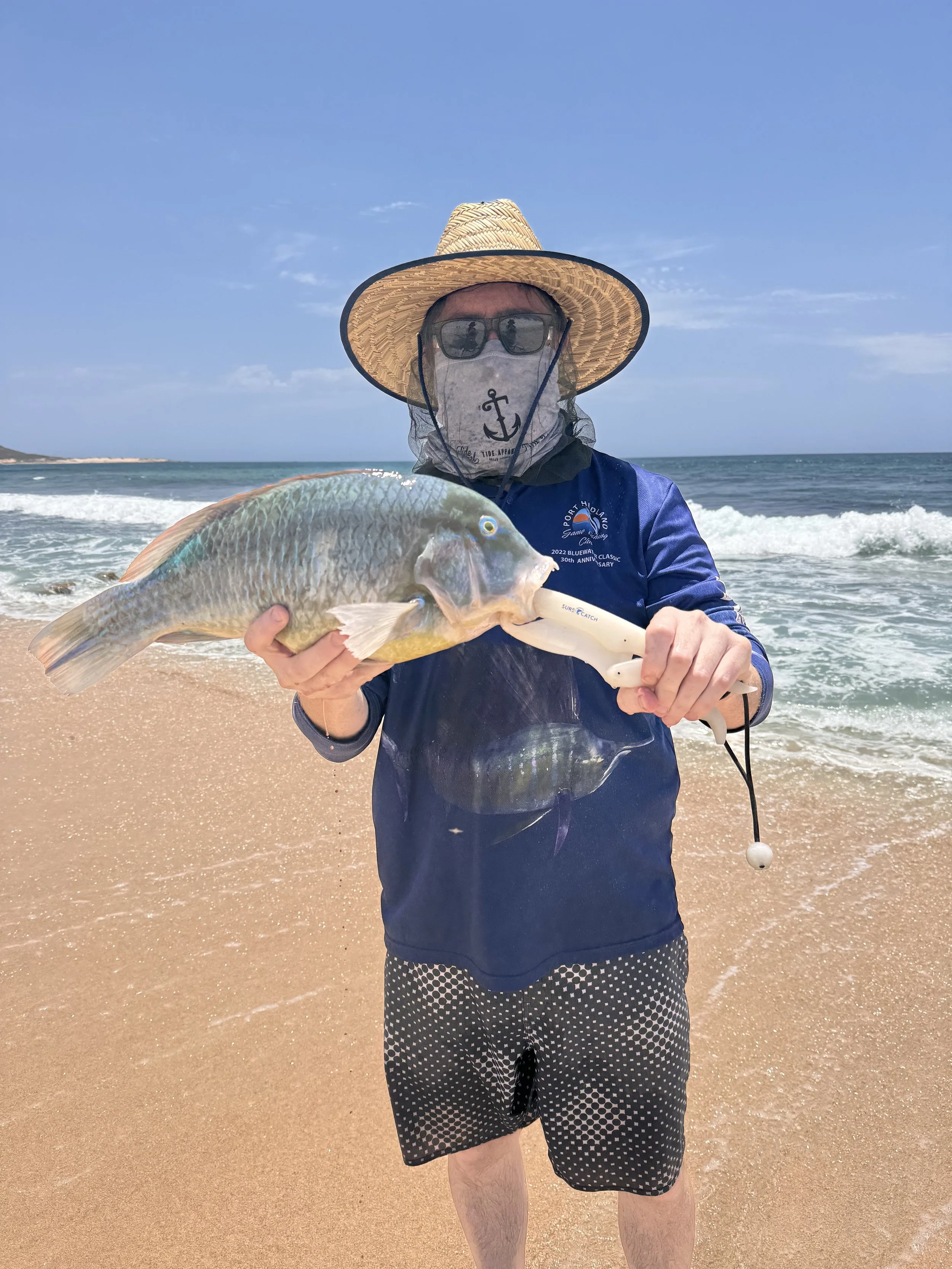 Person holding a fish on a sandy beach with ocean waves in the background, wearing a sun hat, sunglasses, a face mask, a blue jacket, and patterned shorts.