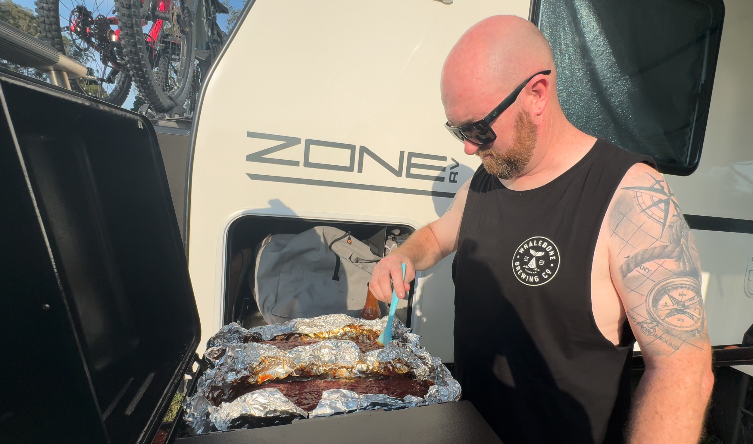 A bald man with tattoos on his arm, wearing sunglasses, standing at a grill with foil-covered food. There are bicycles on a rack behind him, and a vehicle with the words 'ZONE RV' written on it is in the background.