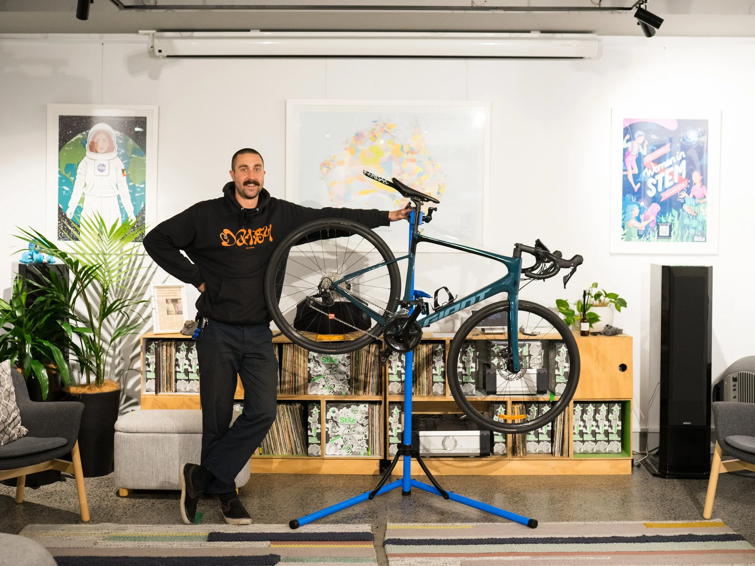 A man standing indoors holding a blue and black road bicycle on a bike stand, smiling at the camera. The background includes framed artwork, a bookshelf, plants, and a black speaker.