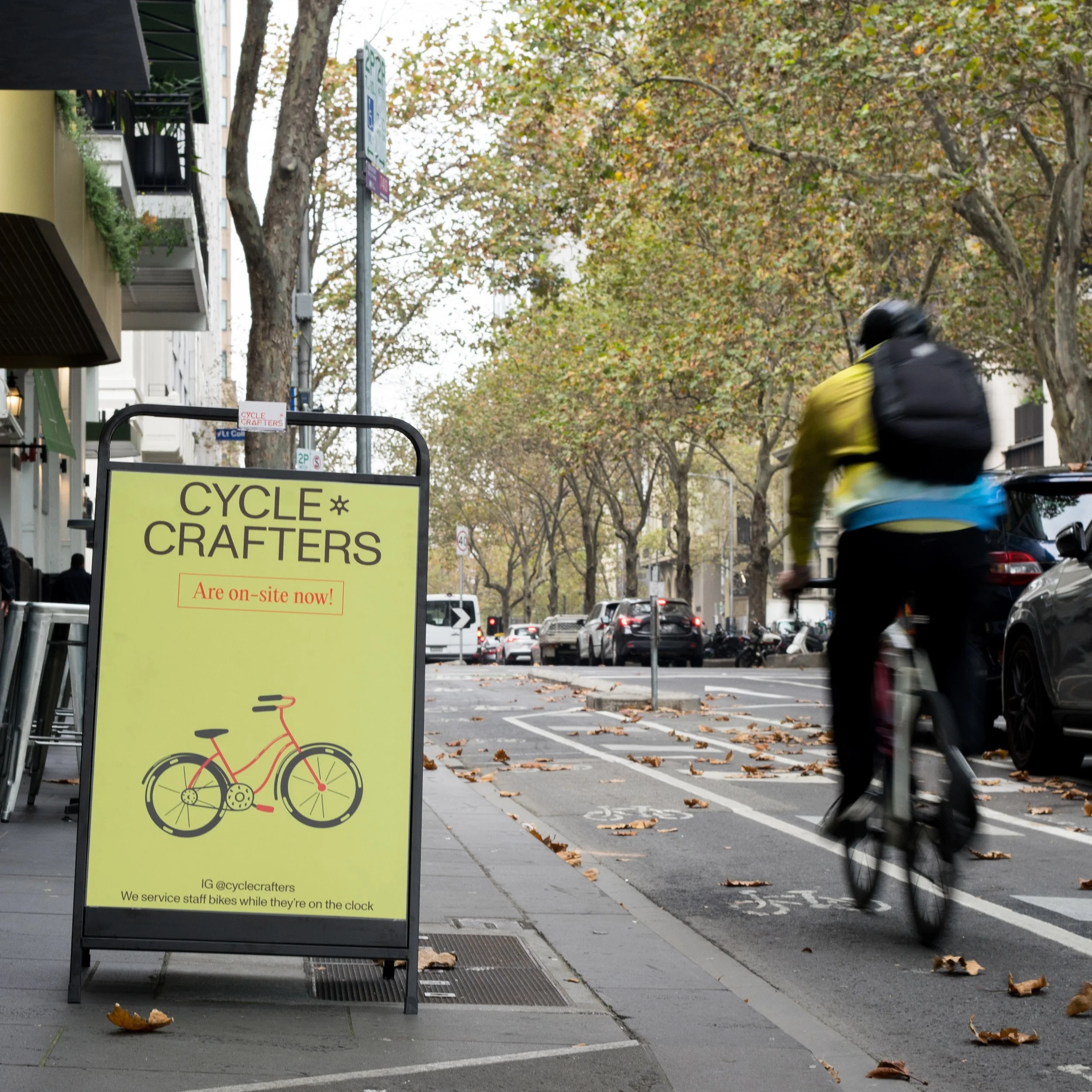 A yellow sidewalk sign for Cycle Crafters on a city street, with a drawing of a bicycle, and a cyclist riding past on a bike lane on the right, lined with trees and parked cars.