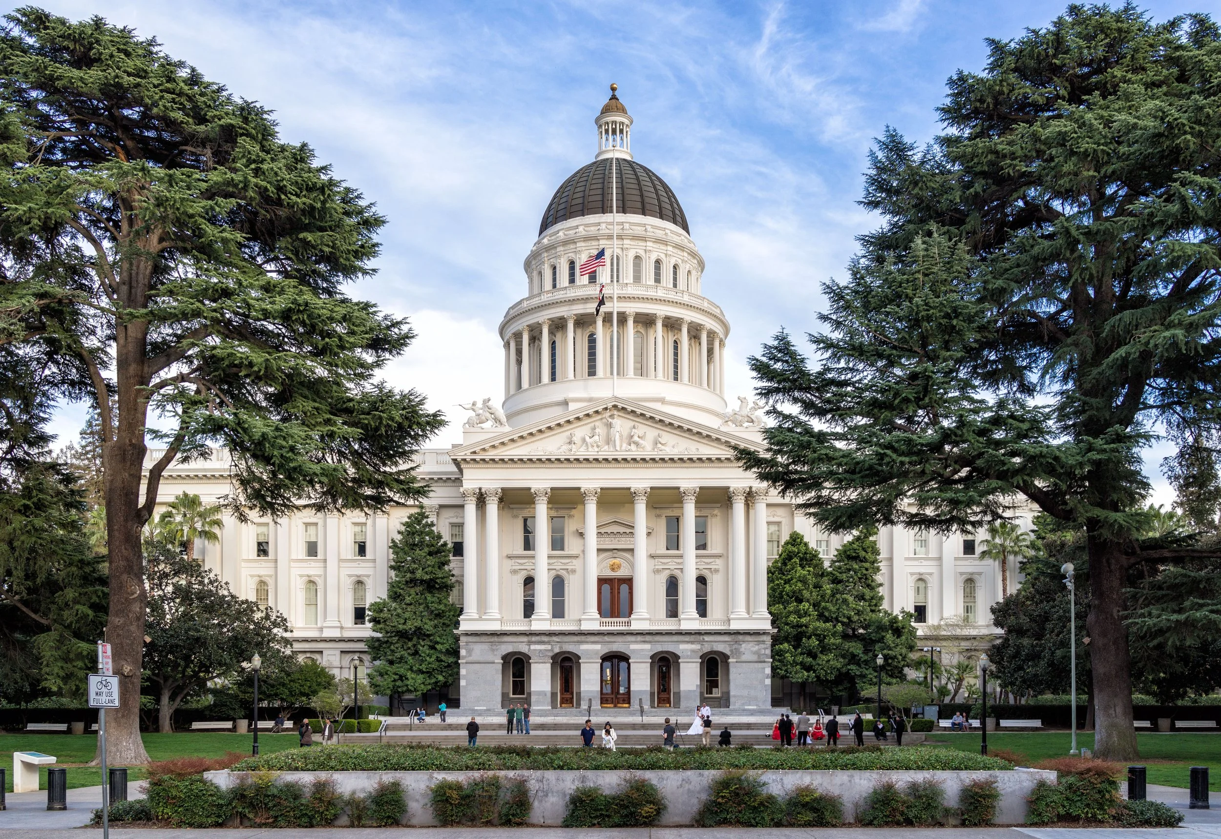 The California State Capitol building in Sacramento, featuring a large dome, white classical architecture, surrounded by trees and people walking in the park.