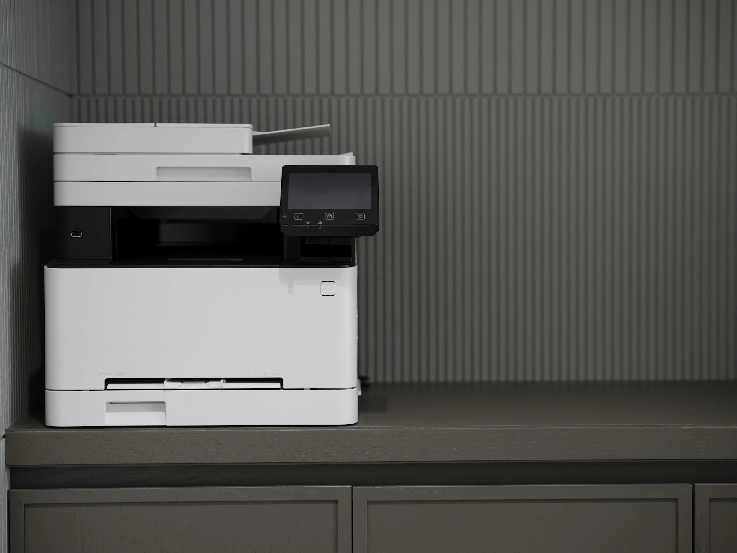 A white multifunctional office printer with a control panel, placed on a dark gray shelf against a gray corrugated wall.