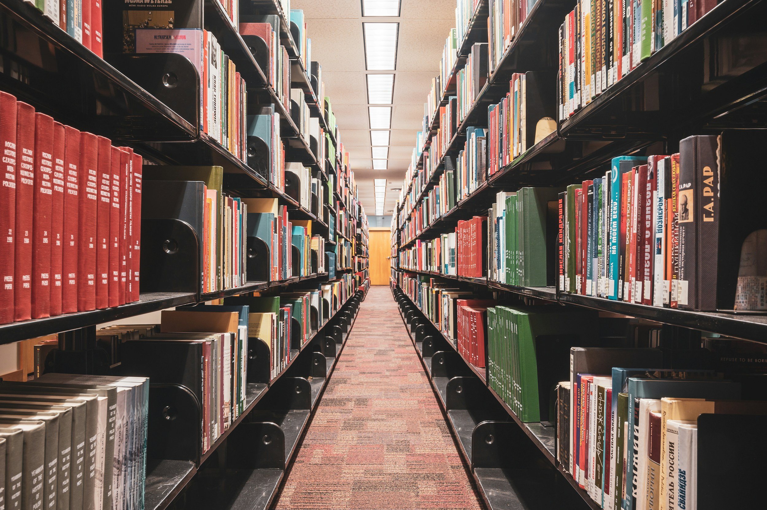 A view down a narrow aisle in a library, with shelves filled with colorful books on both sides and a carpeted floor.