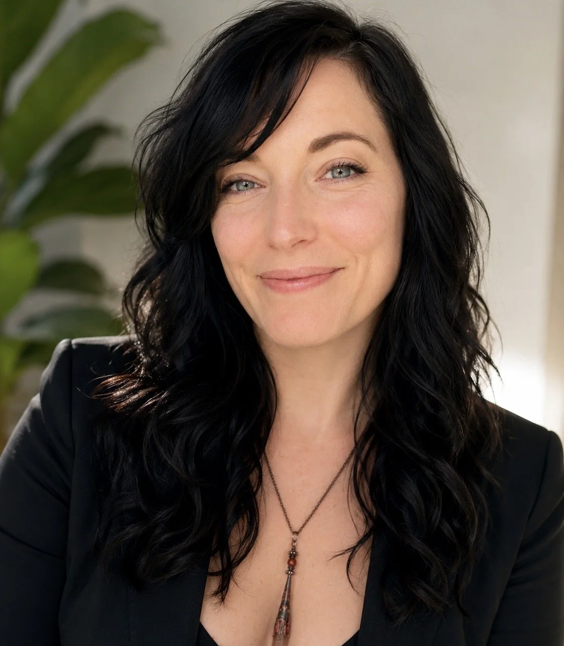 A woman with long, curly black hair, blue eyes, and fair skin smiling, wearing a black blazer and a necklace with a pendant, with a green plant in the background.