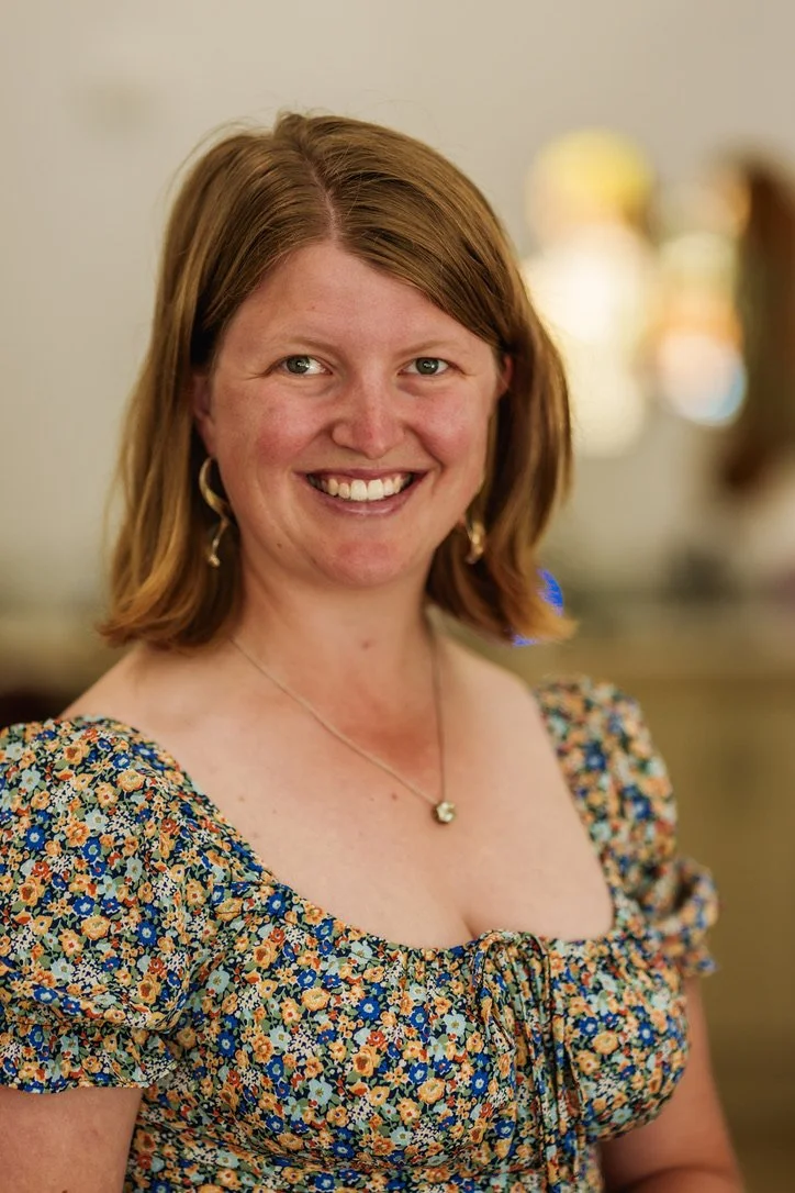 A woman with shoulder-length reddish-brown hair wearing a floral dress with puffed sleeves and a gold heart-shaped necklace, smiling indoors.
