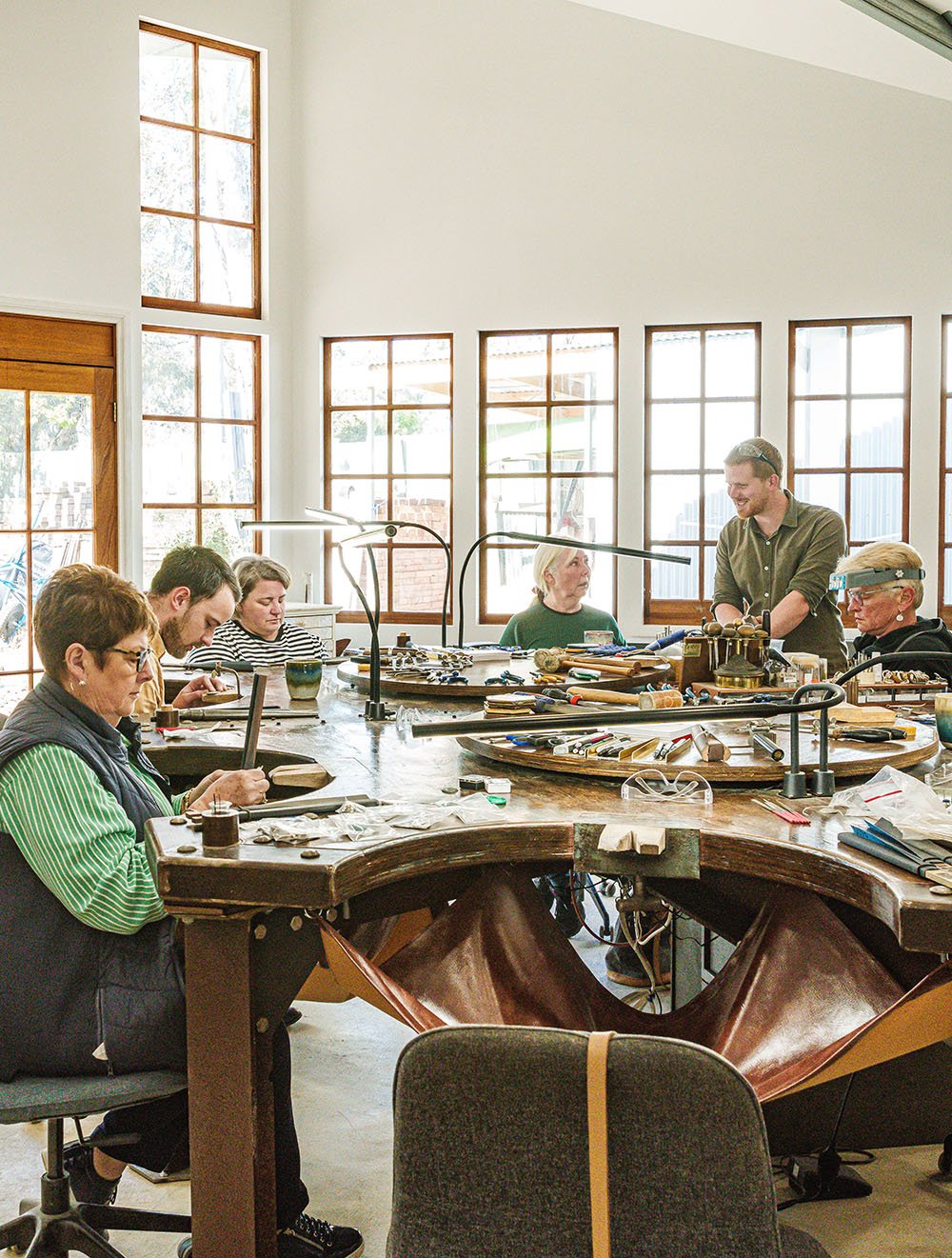 People working on jewelry at a large central workbench in a bright craft room.