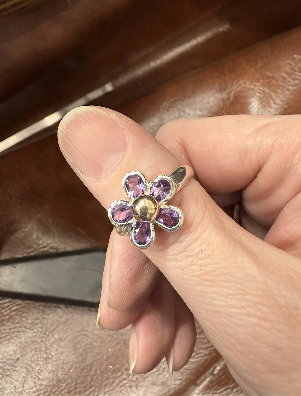 A hand wearing a ring with a flower design featuring purple gemstones and a gold center.