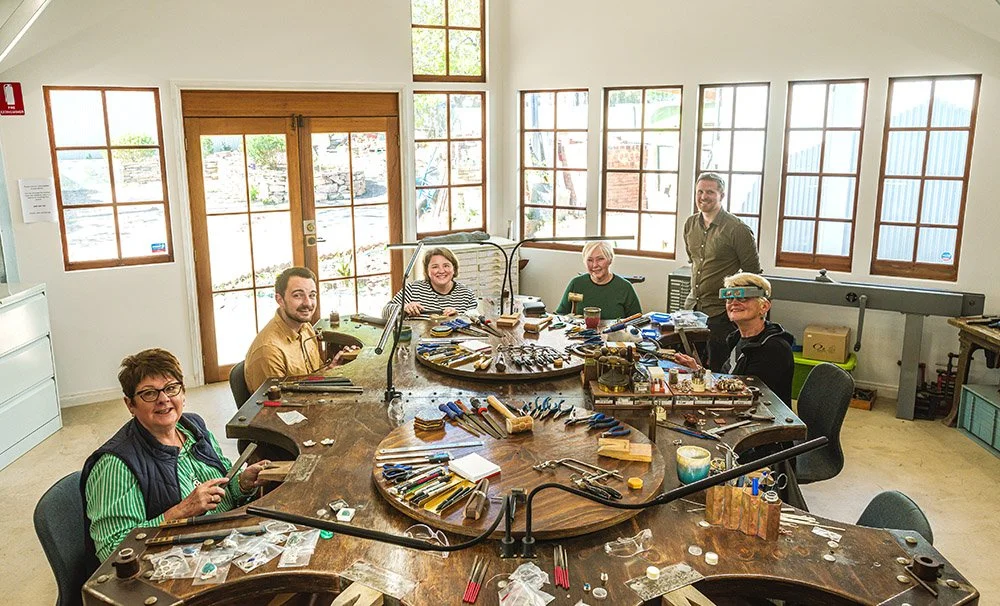 Group of people working at a large, circular woodworking table with tools and materials in a bright workshop.