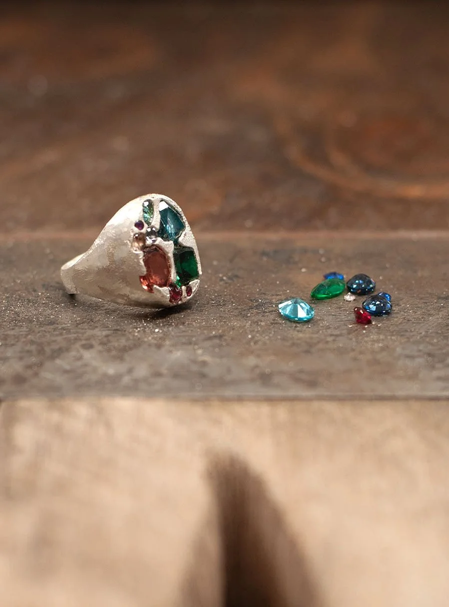 Close-up of a silver ring with colorful gemstones and a few loose gemstones on a wooden surface.