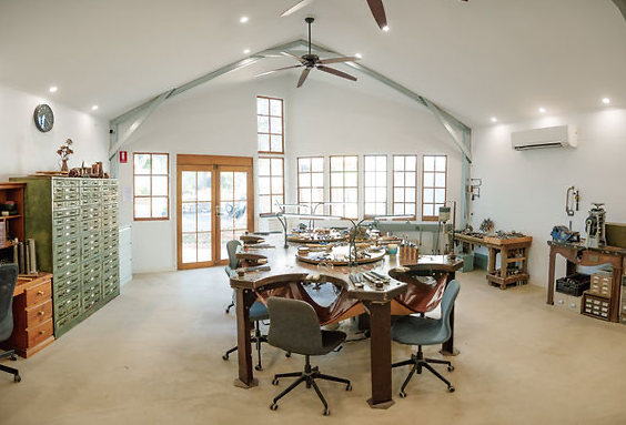 Spacious room with a large circular table set for a meal, surrounded by chairs, set against a background of windows and wooden doors, with shelves and equipment along the walls.