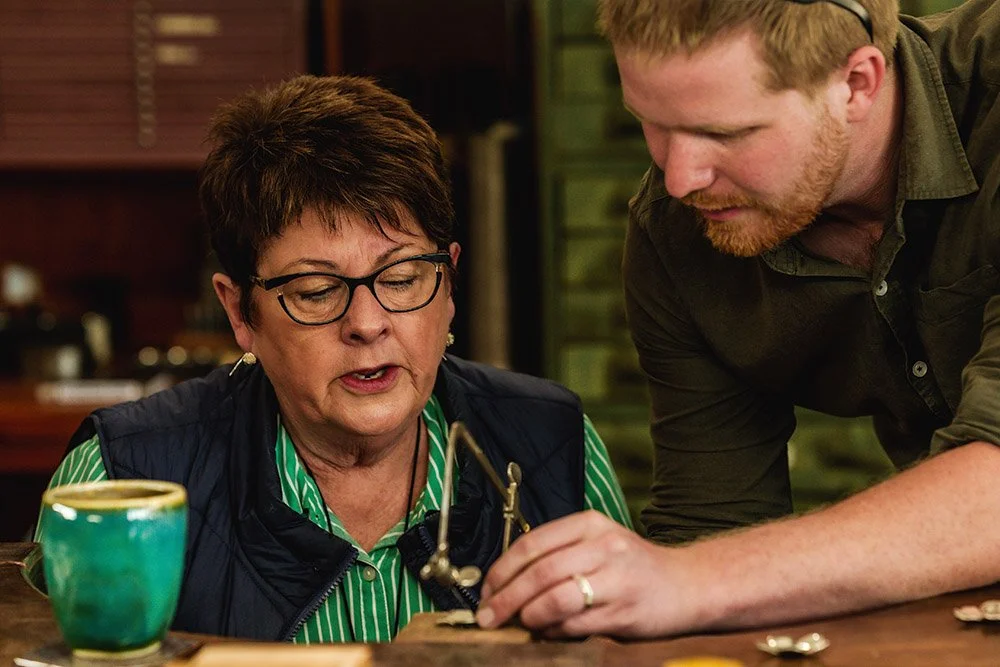 A woman with short dark hair, glasses, and a green striped shirt interacts with a man with red hair and beard, who is leaning over a table. They appear to be engaged in jewellery making in the Adelaide Hills at Hive Workshops.