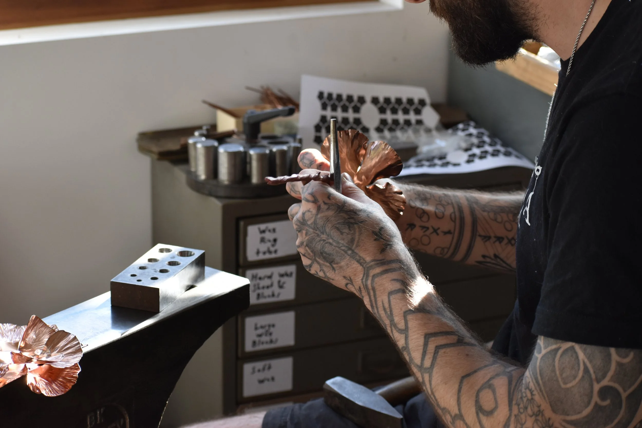 A tattooed man working on a metallic sculpture using a small hand tool in a workshop. Various art supplies and tools are on the table around him, with notes and labels on drawers in the background.