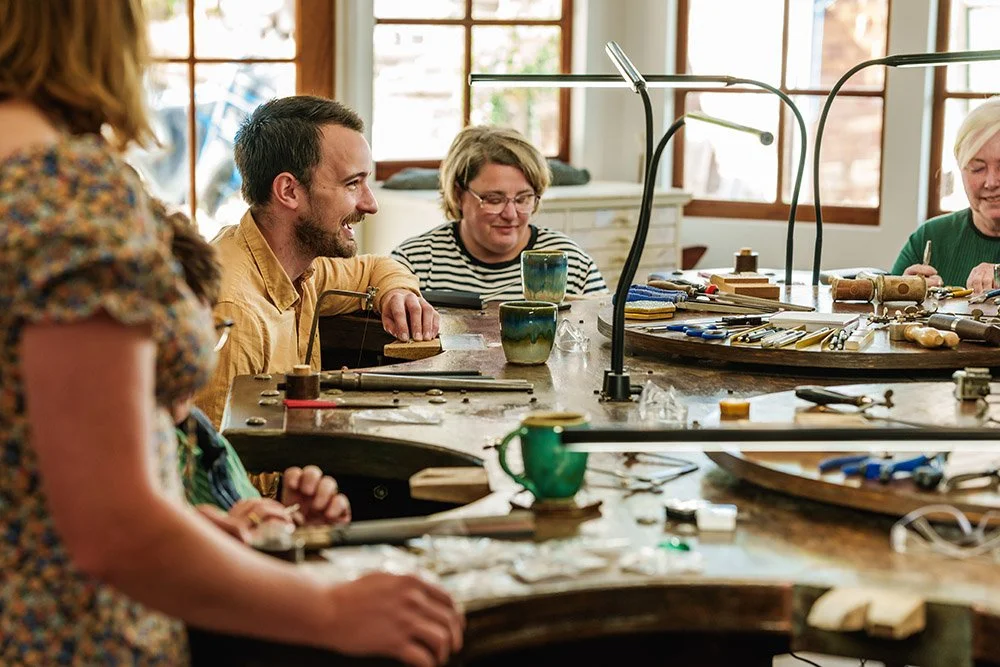 People working on jewelry making at a workshop table with tools, in a well-lit room with large windows.