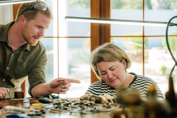 A man and woman are making jewellery a table near a large window in a bright room.