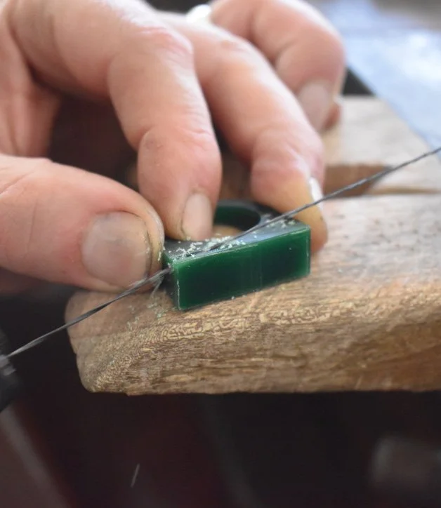 A person's hand holding a small green block or object against a wooden surface, with a thin wire passing over it, in a woodworking or crafting context.