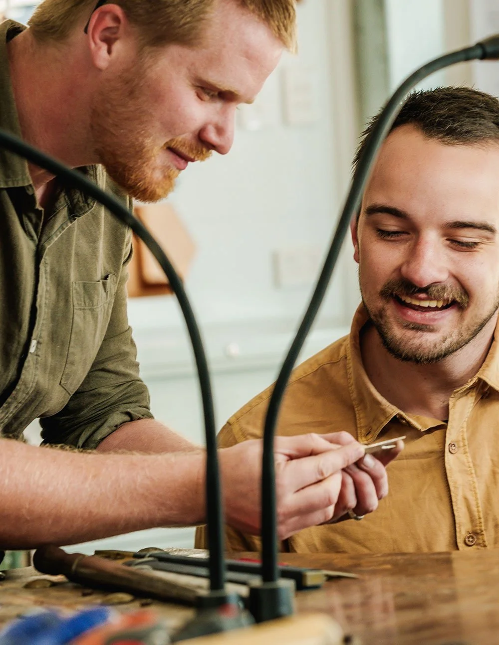Two men smiling and looking at a silver ring they are making, with jewellery mkaing equipment on the table in front of them.