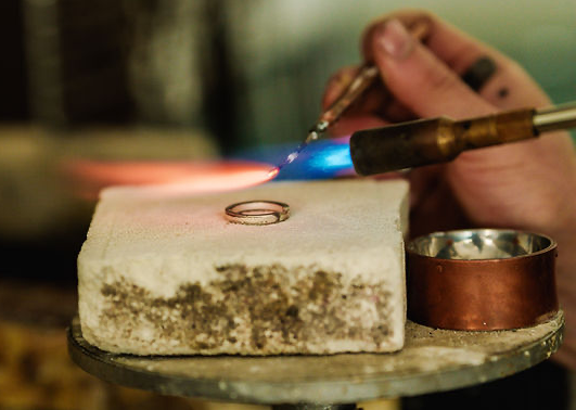 A person is melting a gold ring with a torch on a small cement block, with a metal container nearby.