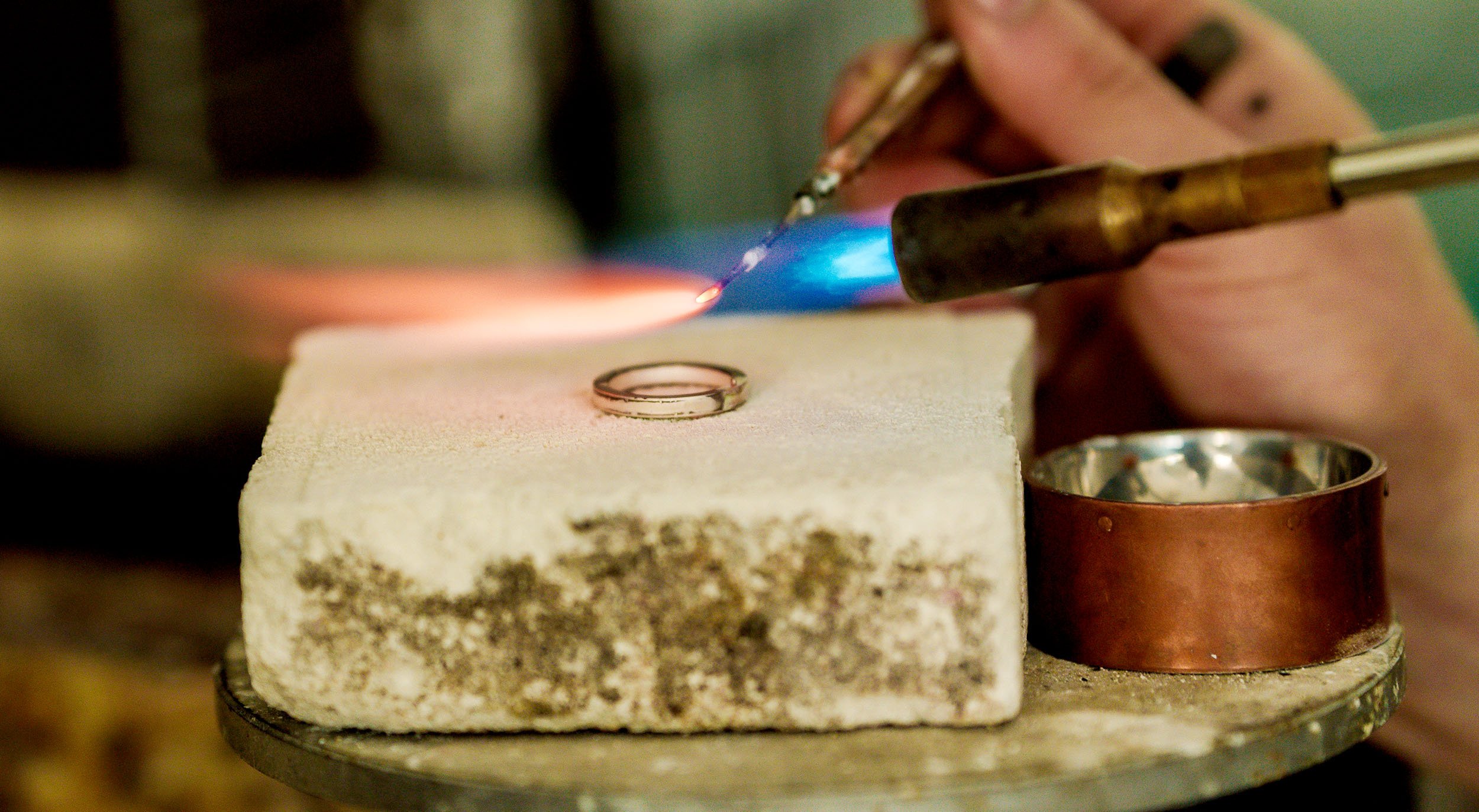 A jeweler is using a torch with a blue flame to work on a metal ring, which is placed on a white stone. The jeweler's hands are visible, holding the ring and the torch.