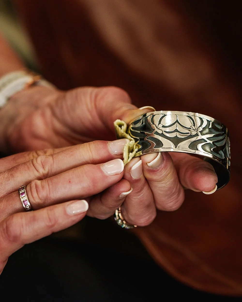 Close-up of hands holding a silver bracelet, with one hand pulling apart a part of the bracelet. The hands have rings, and the background is blurred.