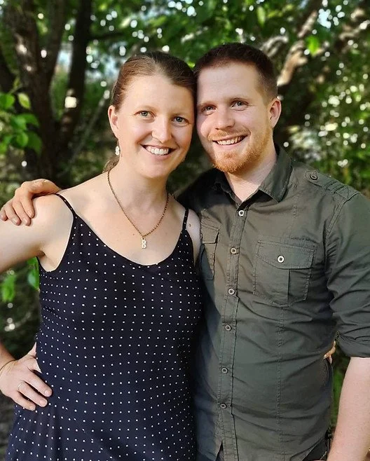 A smiling woman and man standing outdoors with green trees in the background, close together, with the woman's arm around the man's shoulder.