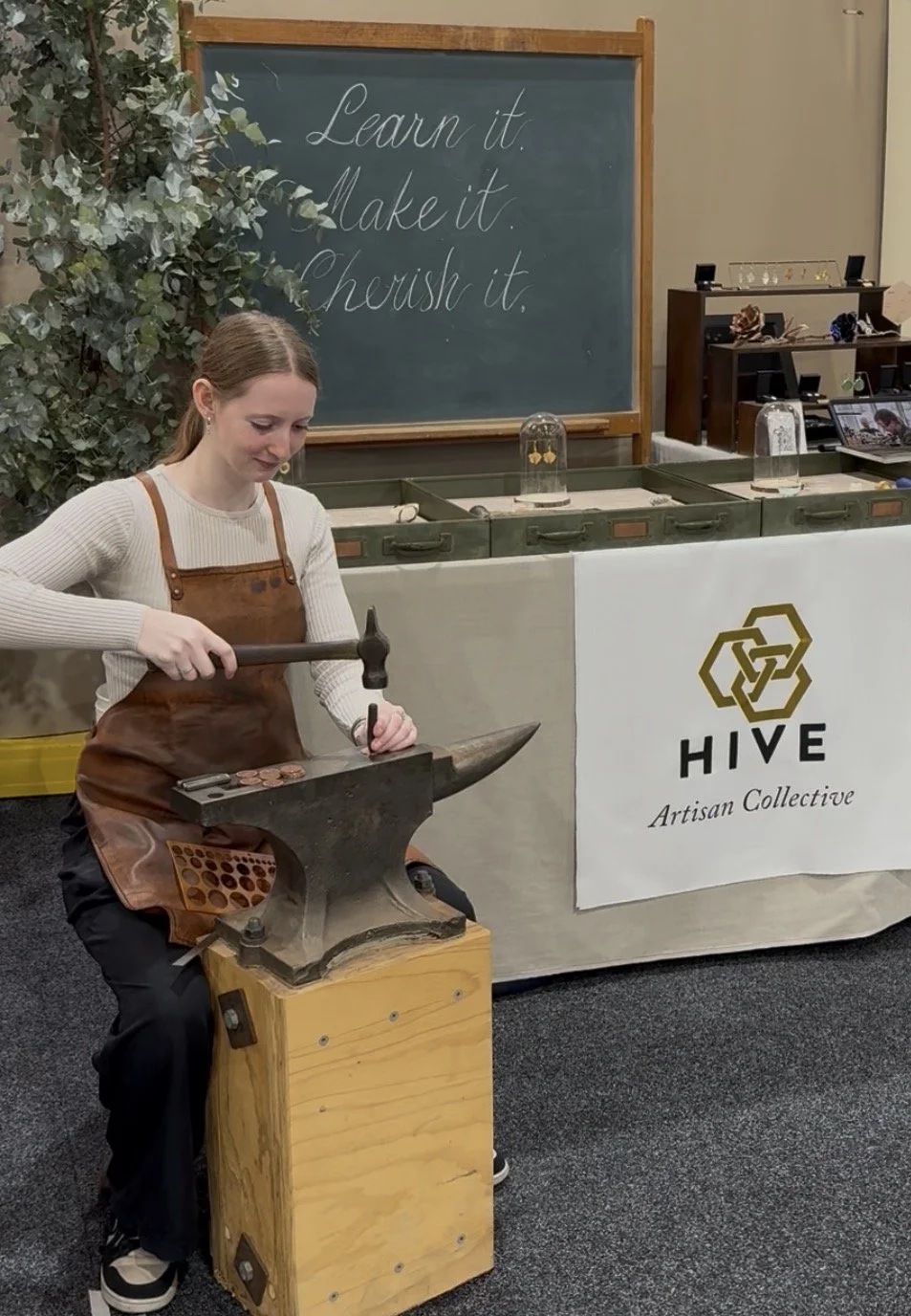 A woman wearing a white sweater and brown apron hammering a metal coin into an anvil at a booth labeled "HIVE Artisan Collective." A blackboard behind her displays the message "Learn it. Make it. Cherish it."