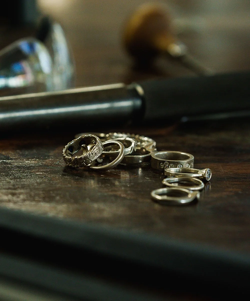 A collection of silver jewelry, including rings and earrings, scattered on a wooden surface with tools and a broken glass in the background.