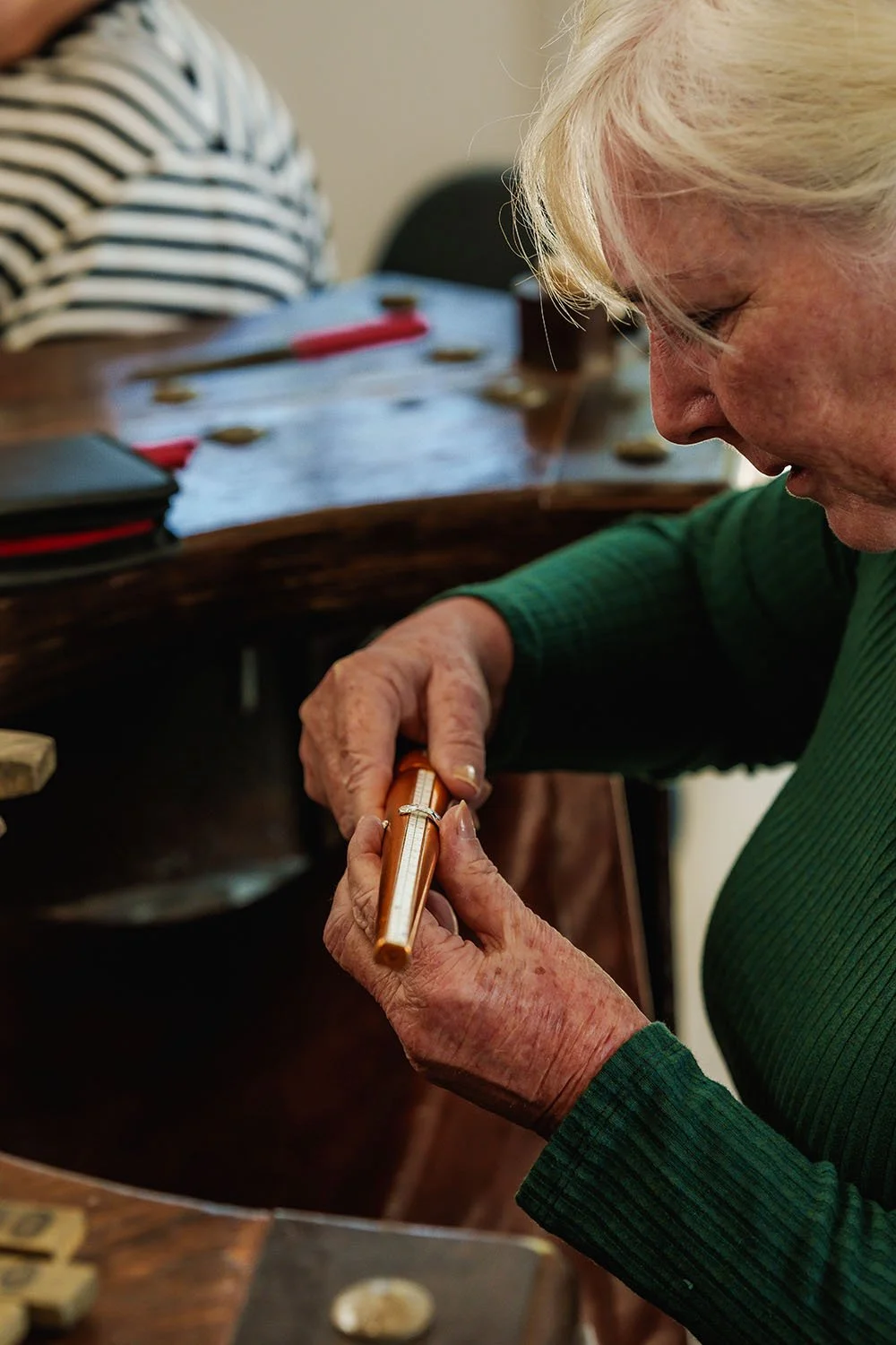 An elderly woman with white hair playing a game of bingo, holding a bingo marker near her mouth, at a table with bingo cards and tokens.