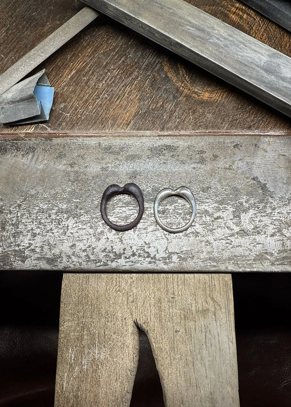 Two rings, one black and one silver, placed on a textured wooden surface. Other jewellery tools and materials are visible in the background.