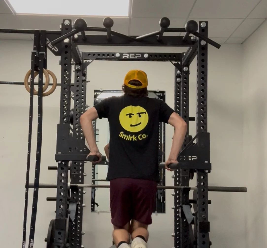Person with long hair wearing a yellow cap, black Smirk Co. t-shirt, and maroon shorts doing a dip exercise on a power rack in a gym.