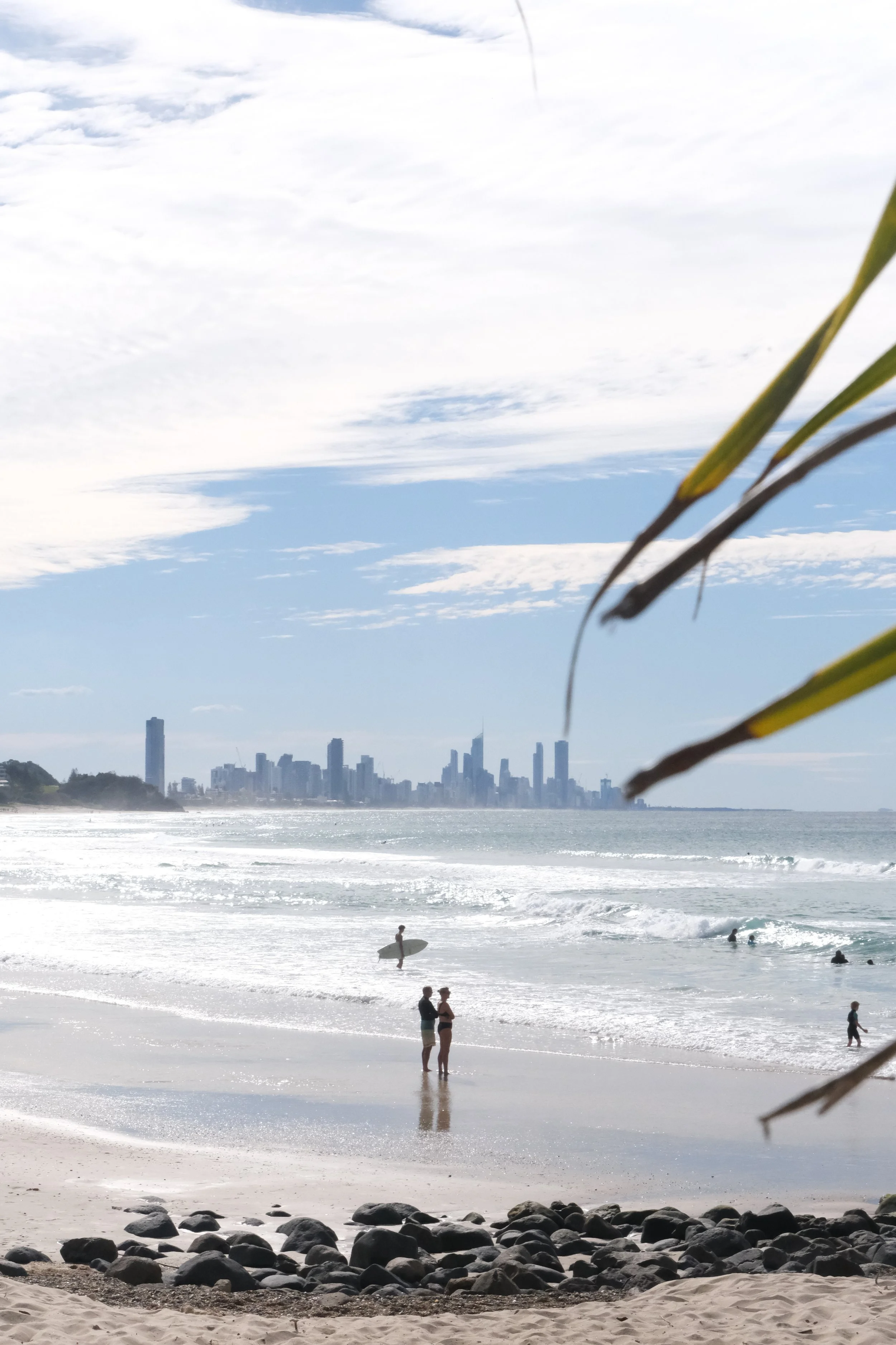 People at the beach with the skyline of a city in the background, surfboards, and some rocks in the foreground.