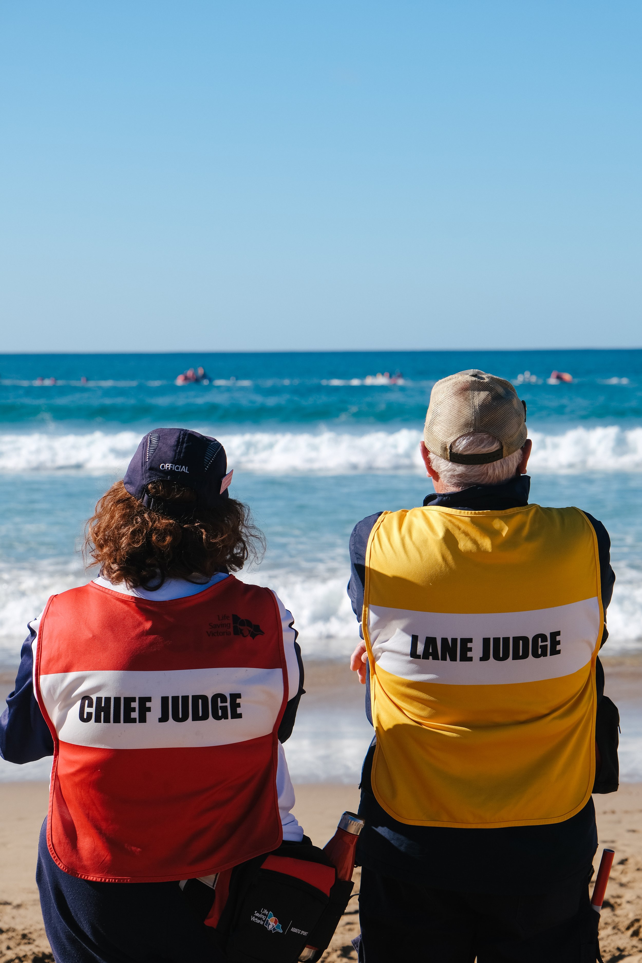 Two lifeguards, one with a red vest labeled 'Chief Judge' and the other with a yellow vest labeled 'Lane Judge', sitting on the beach and observing the ocean with swimmers in the water.