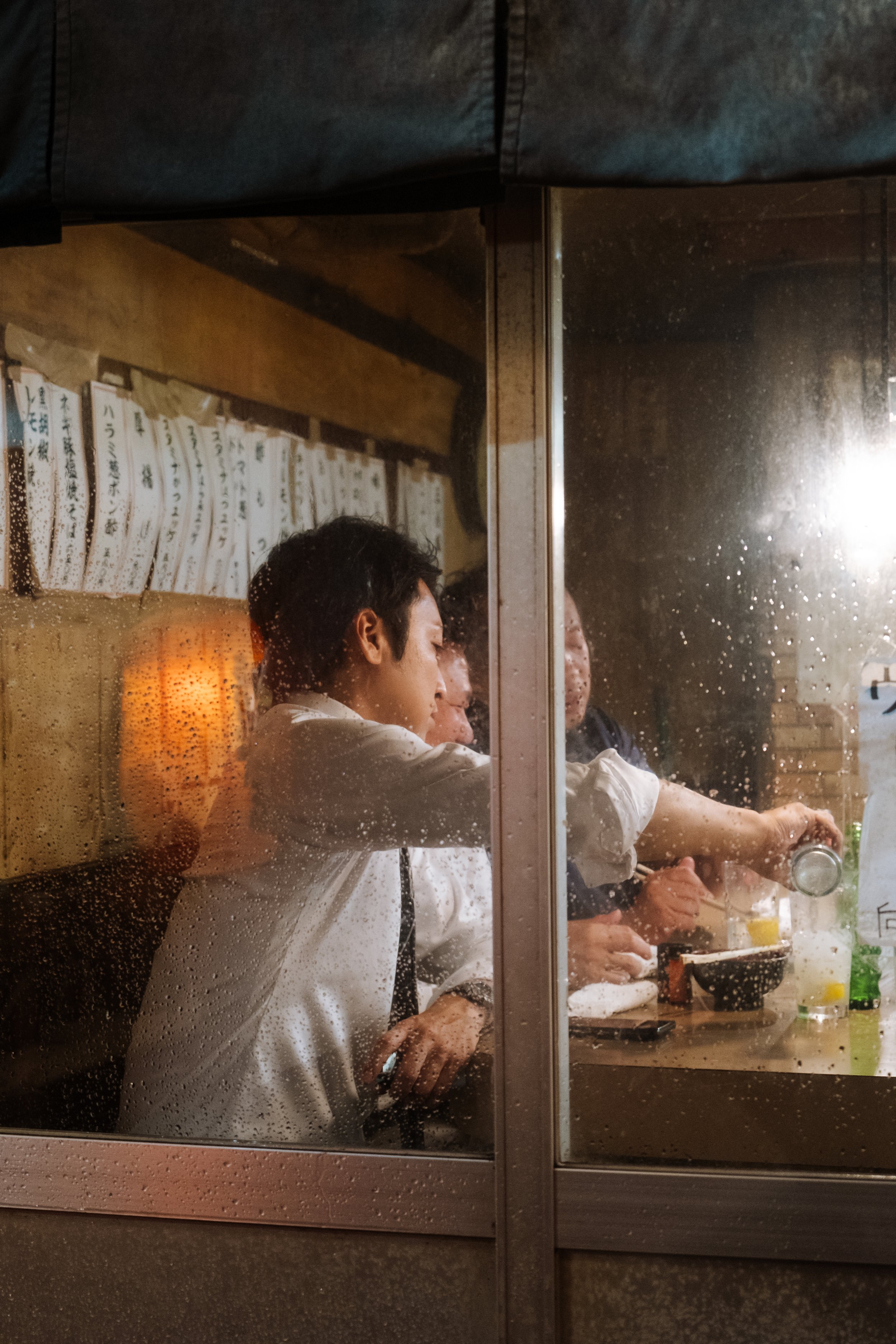 People dining inside a restaurant viewed through rainy glass window, with some condensation on the glass, in a cozy setting.
