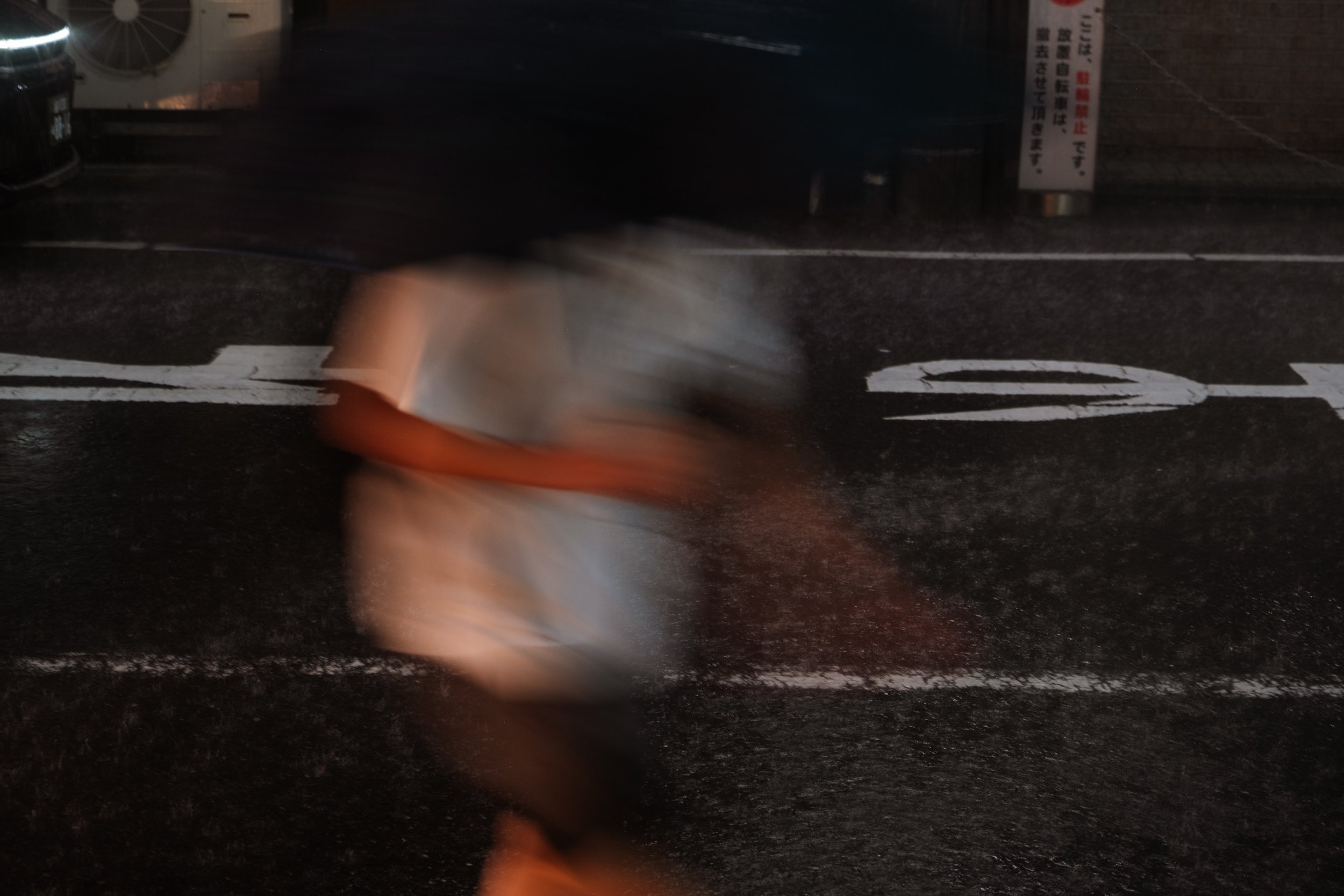 Blurred motion of a person walking across a crosswalk at night, with street markings and signs visible in the background.