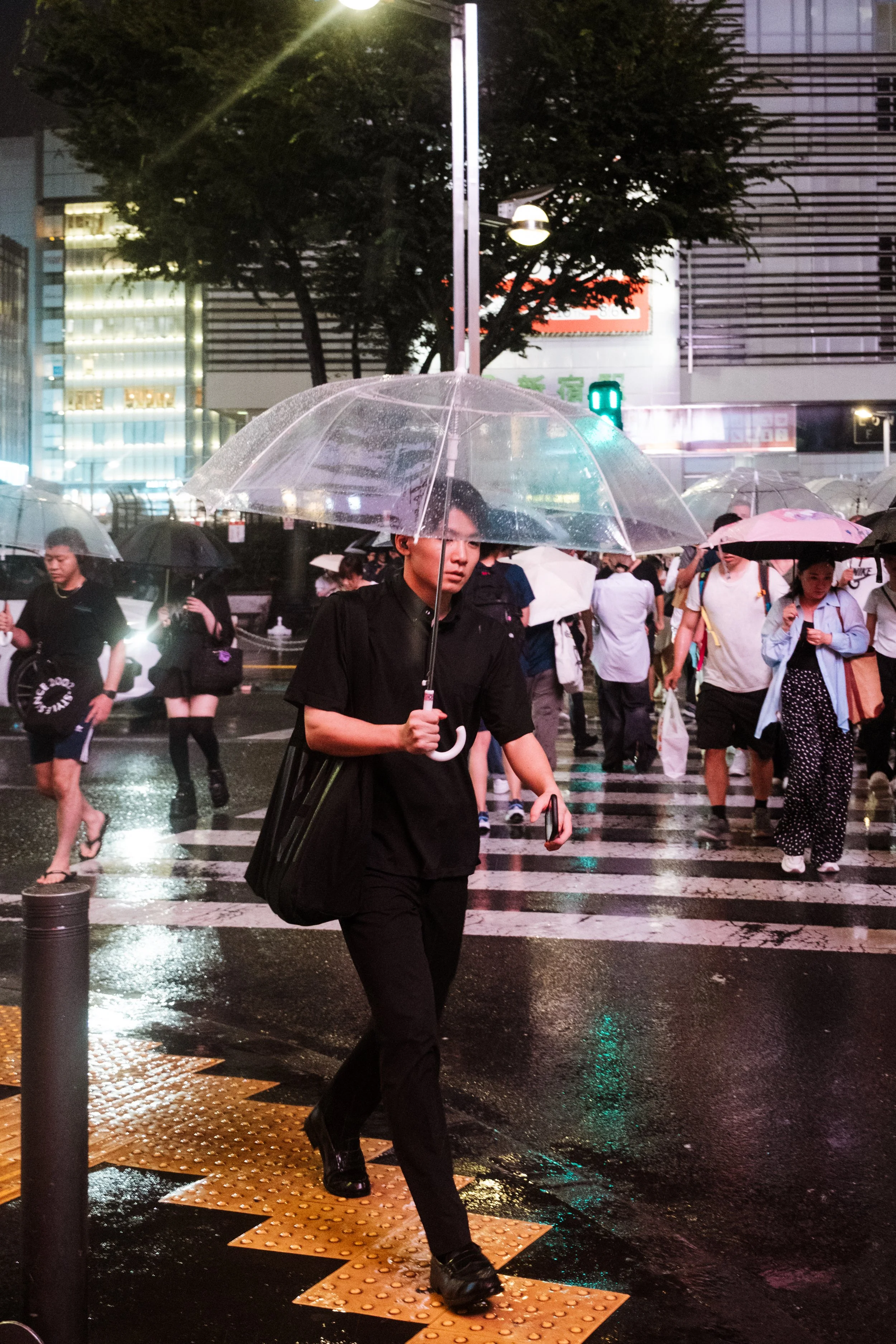 People crossing a city street at night during rainy weather, holding umbrellas, with wet pavement reflecting city lights, and tall buildings in the background.