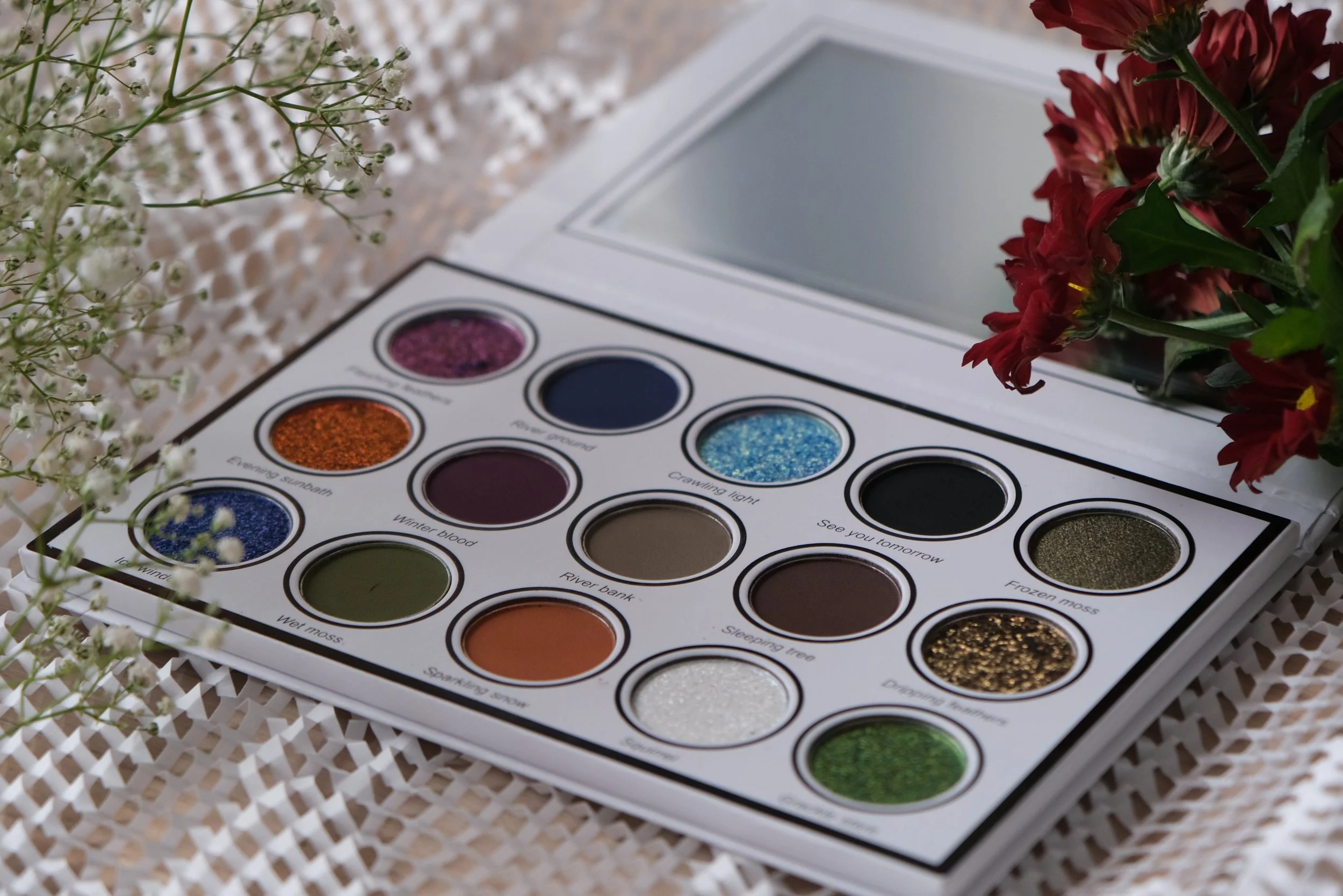 An open makeup palette with 15 eyeshadow colors, surrounded by white baby's breath and red flowers, on a white woven surface.