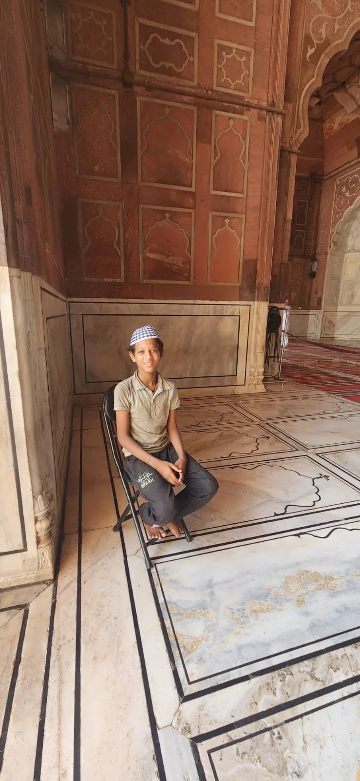 A young boy sitting on a black metal chair inside a historical building with intricate marble flooring and ornate red and beige walls.