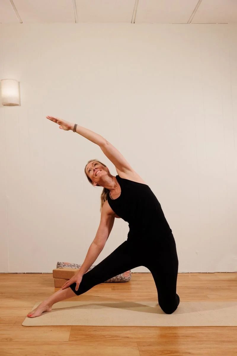Woman practicing yoga on a mat, kneeling with one leg extended to the side and stretching her arm overhead in a yoga pose.