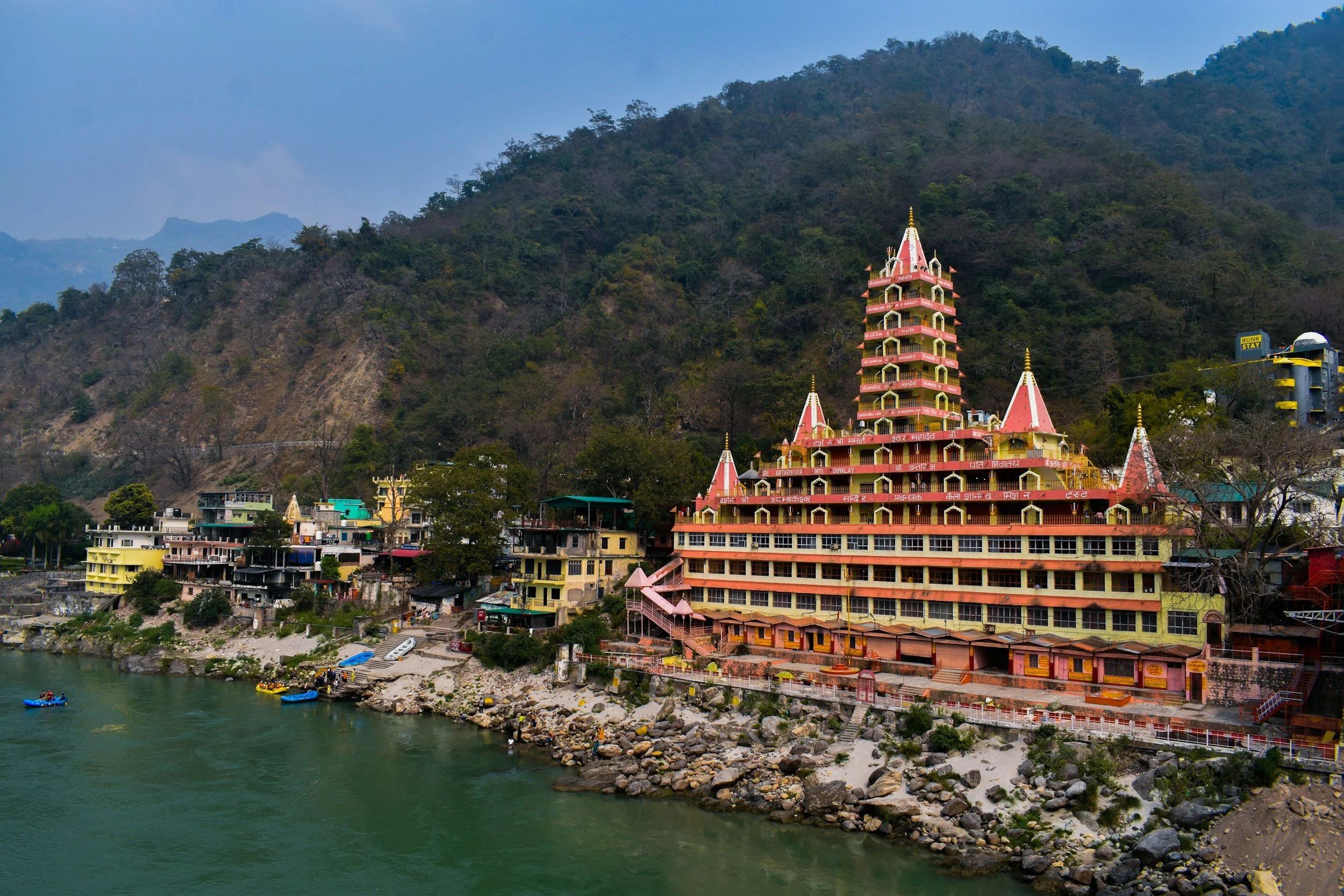 Colorful hilltop temple beside river, with small buildings and forested mountains in background.